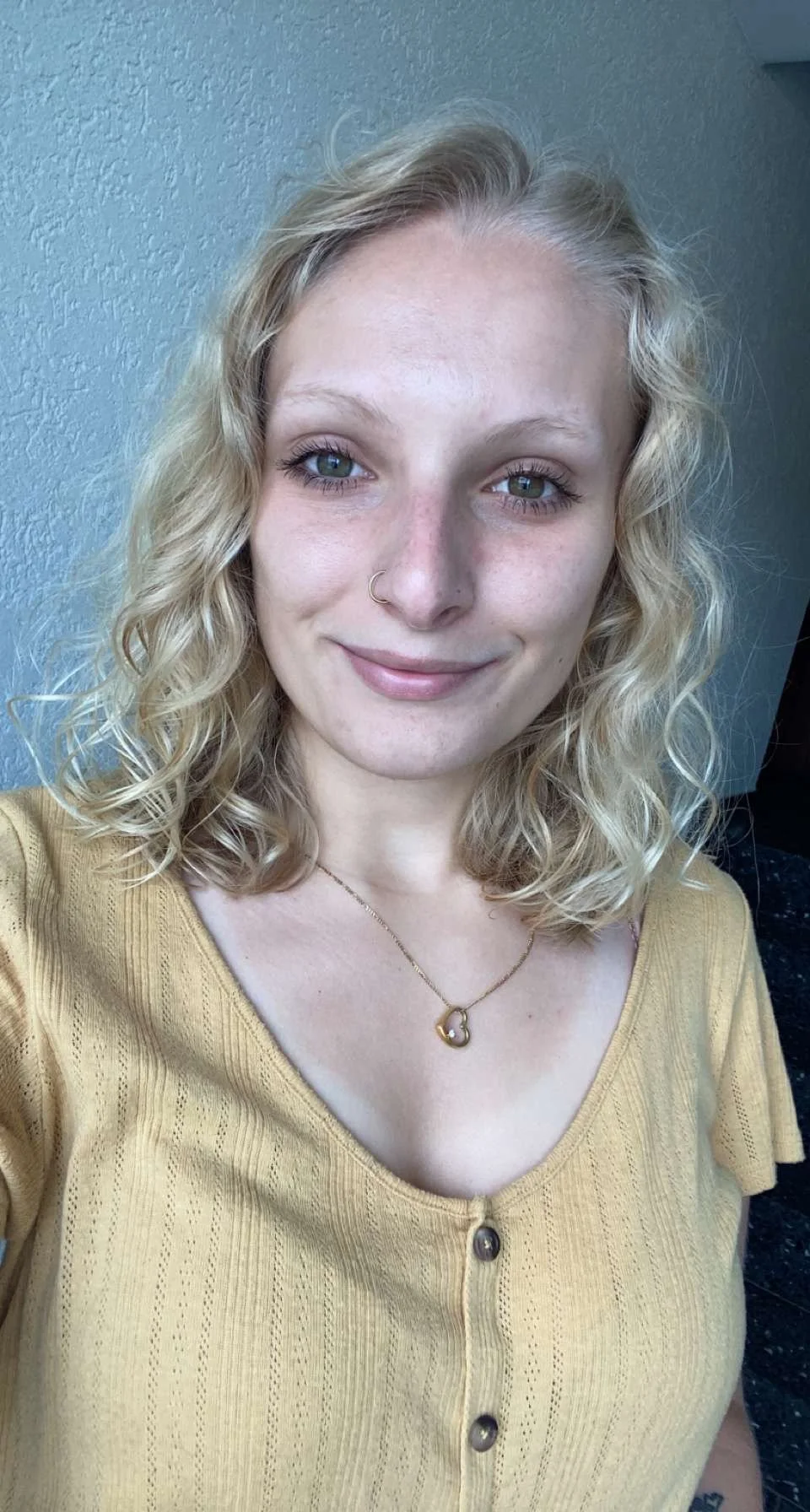 A young woman with blonde curly hair, blue eyes, and fair skin taking a selfie indoors. She is wearing a mustard yellow top with buttons and a gold necklace with a heart-shaped pendant.
