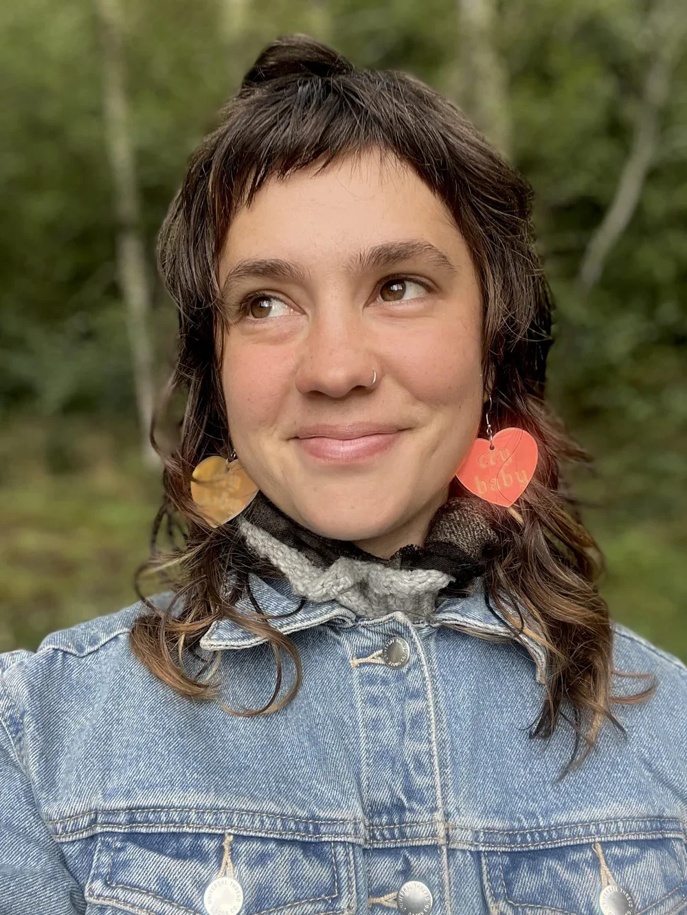 Close-up of a smiling young woman outdoors, wearing hoop earrings with heart-shaped charms, a nose ring, a denim jacket, and a scarf, with a blurred green forest background.