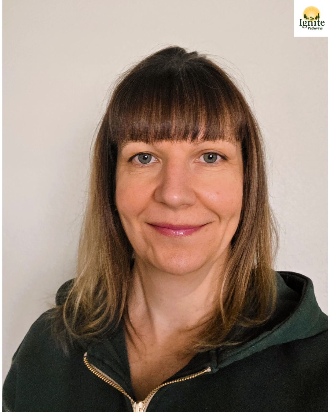 A woman with shoulder-length brown hair and bangs smiling at the camera, wearing a black zip-up jacket, with a blank white wall in the background.