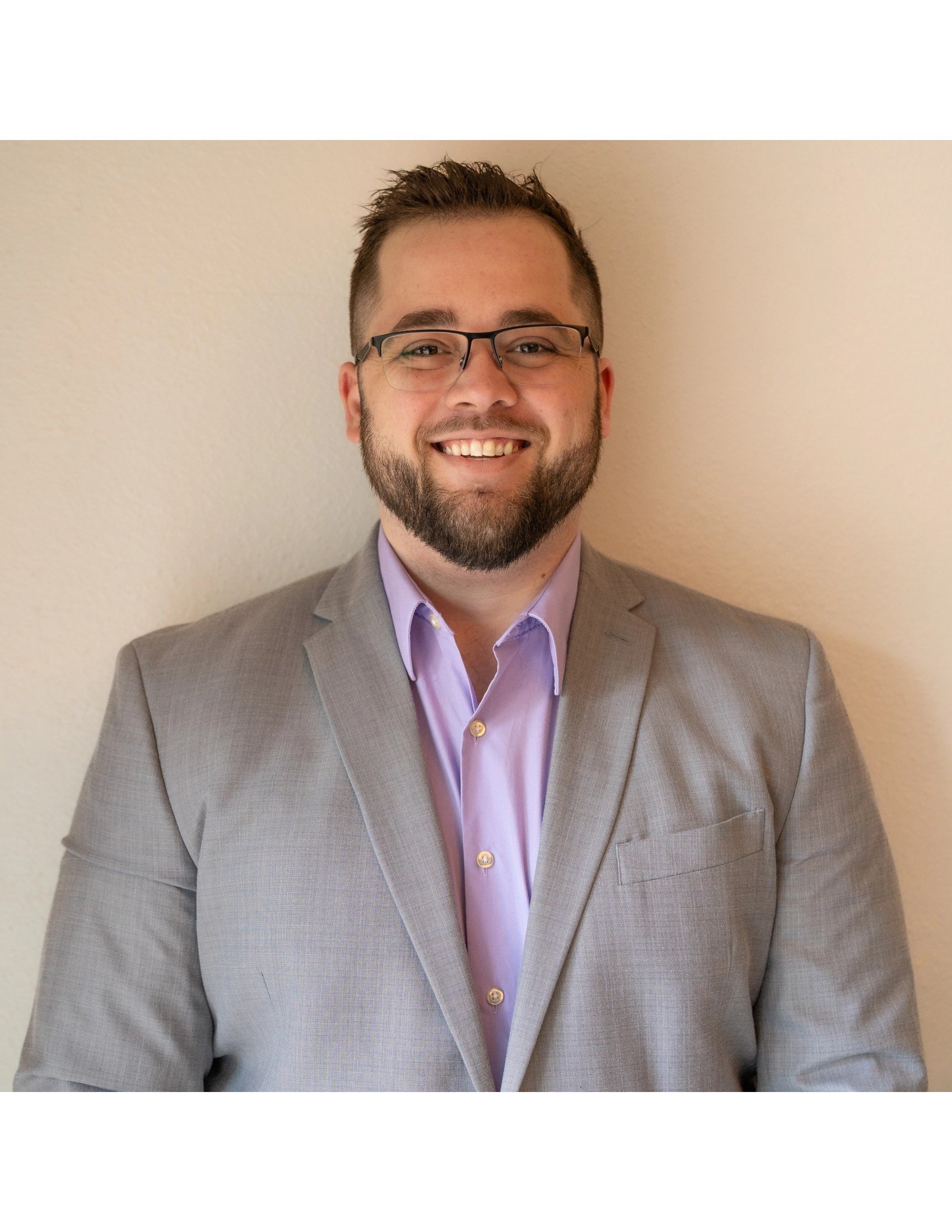 A smiling man with glasses and a beard with brown hair, wearing a light purple shirt and a gray suit, standing against a beige wall.