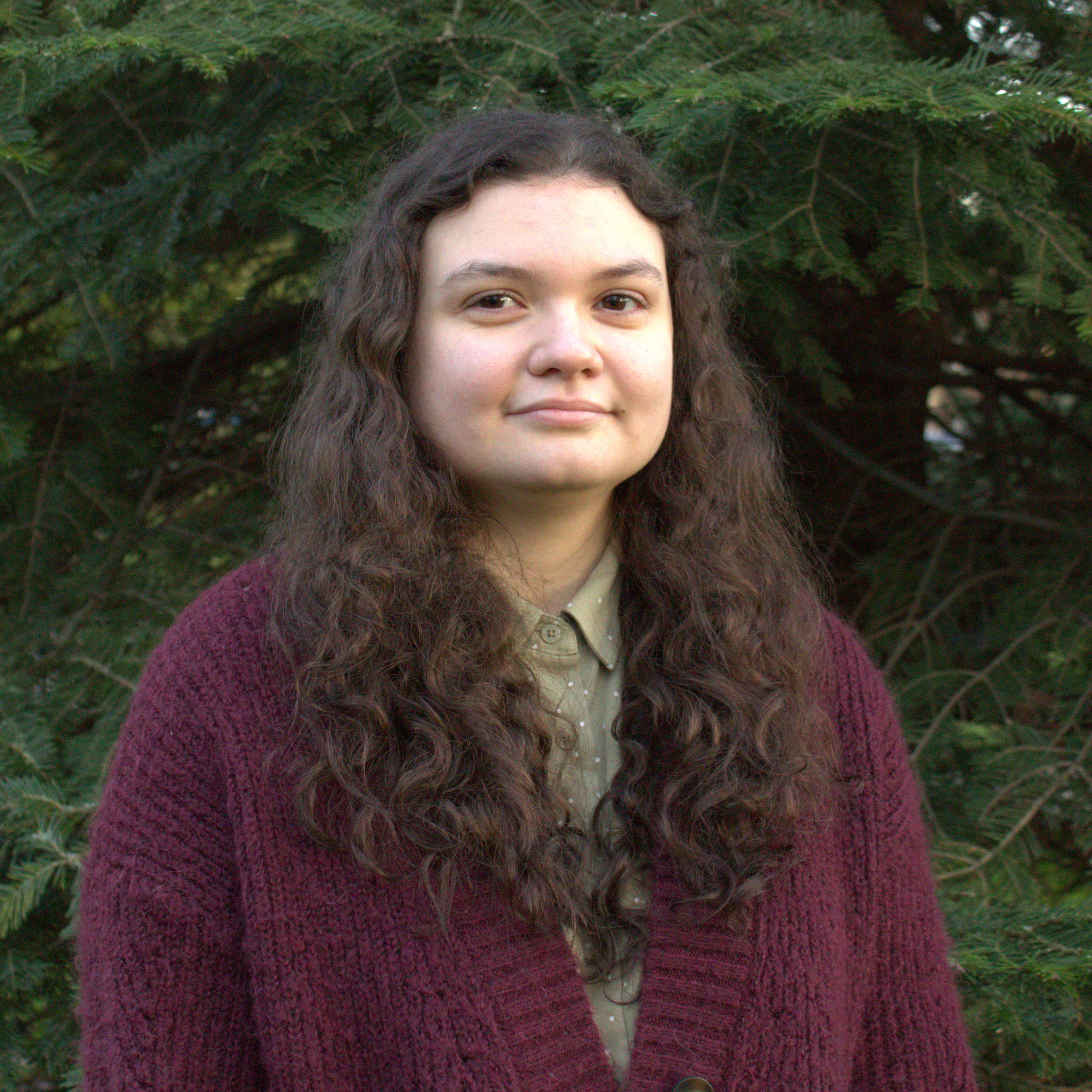 Close-up of a woman with long, wavy hair and fair skin, standing outdoors in front of green pine trees, wearing a beige top and a maroon knitted cardigan.