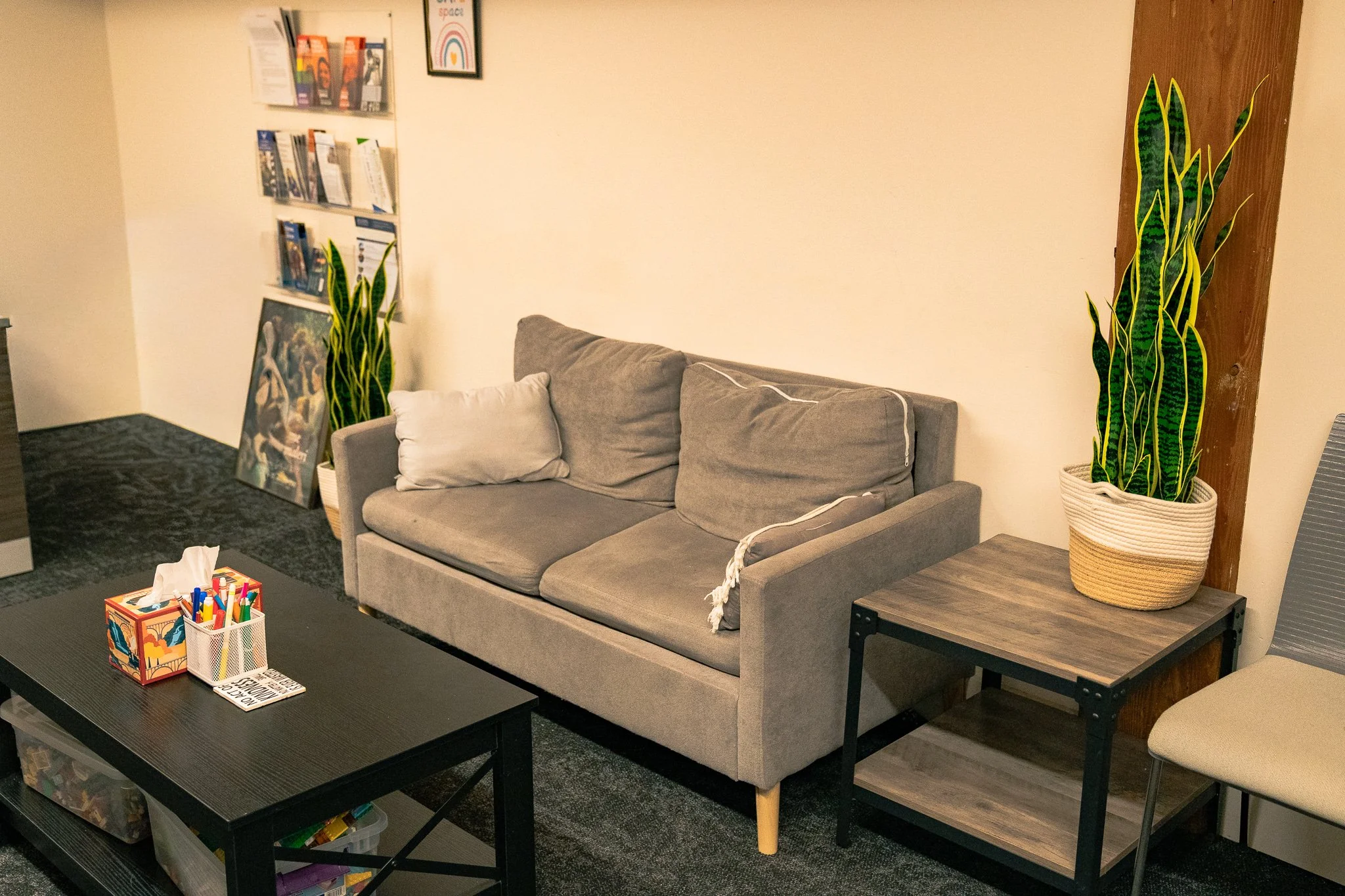 A cozy room corner with a gray couch, decorative pillows, potted snake plants, a black coffee table with art supplies, a bookshelf with magazines, and a framed rainbow artwork on the wall.