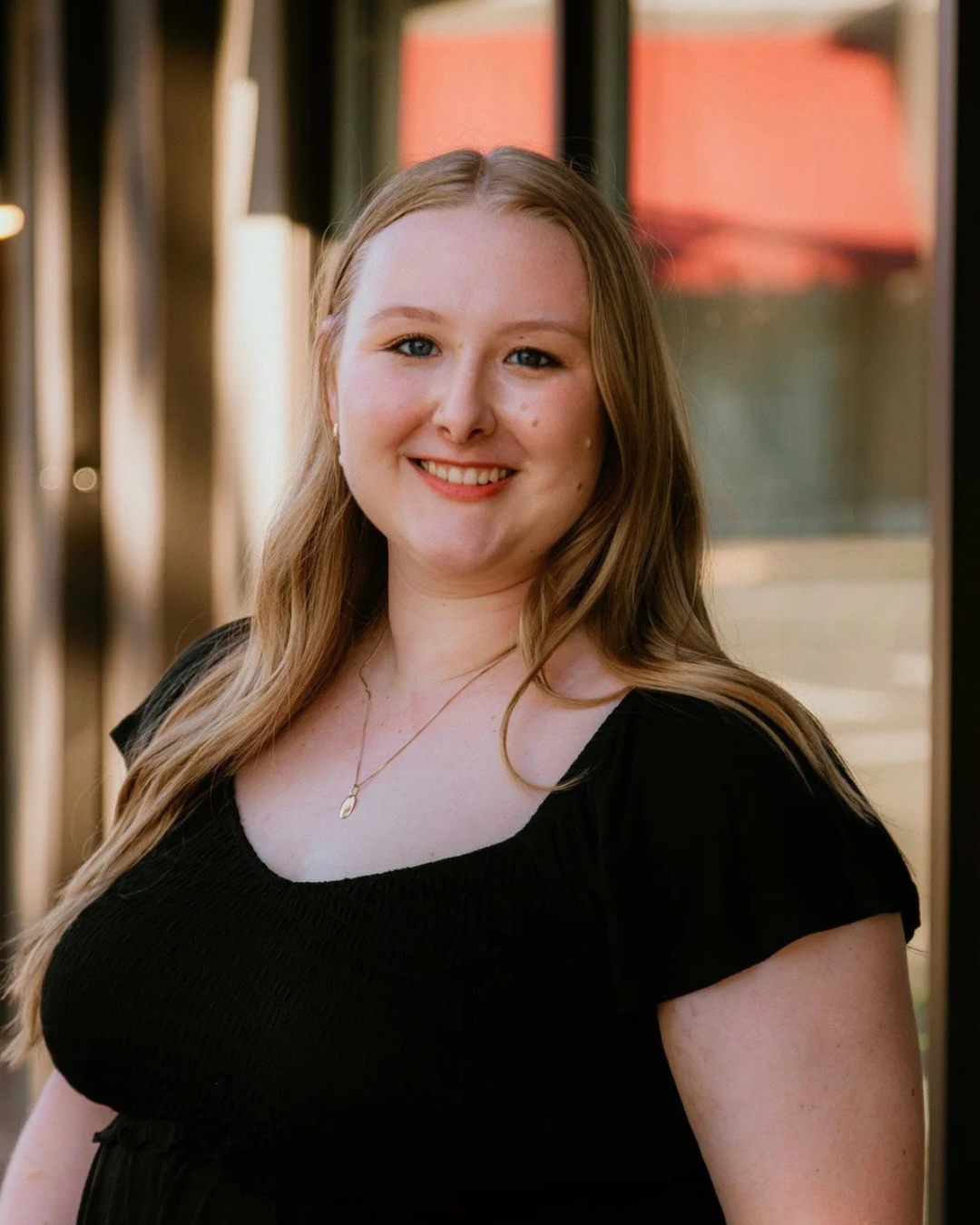A young woman with long blonde hair smiling outdoors, wearing a black top and a gold necklace, with a background of glass windows and a blurred building.