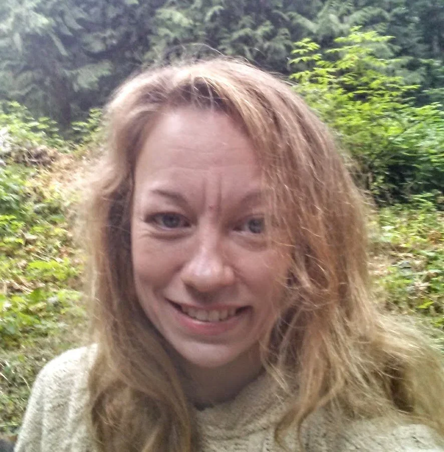 A woman with wavy red hair smiling in a lush, green outdoor setting.