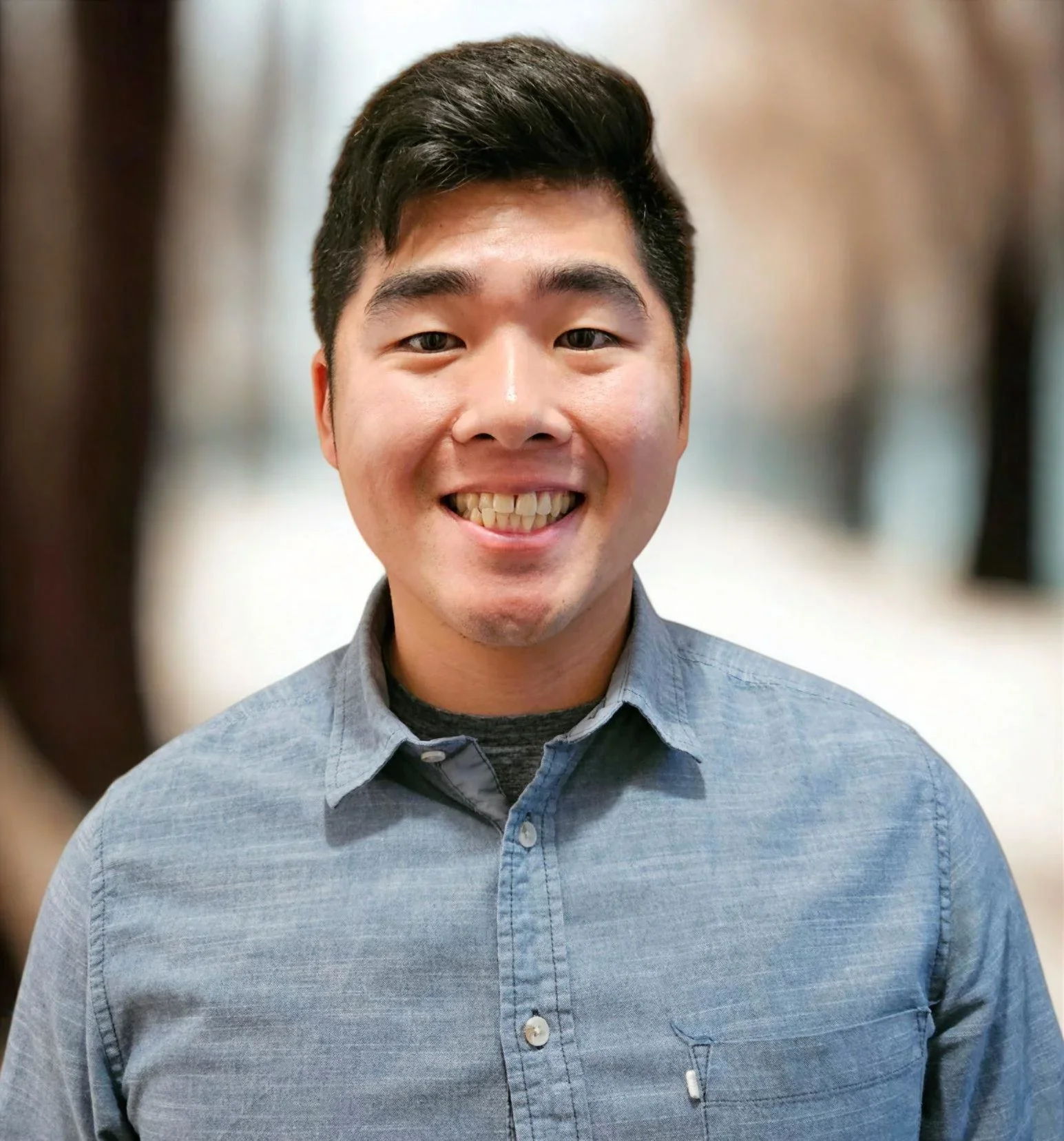A young man with dark hair smiling, wearing a light blue button-up shirt and a dark undershirt, standing indoors with a blurred background.