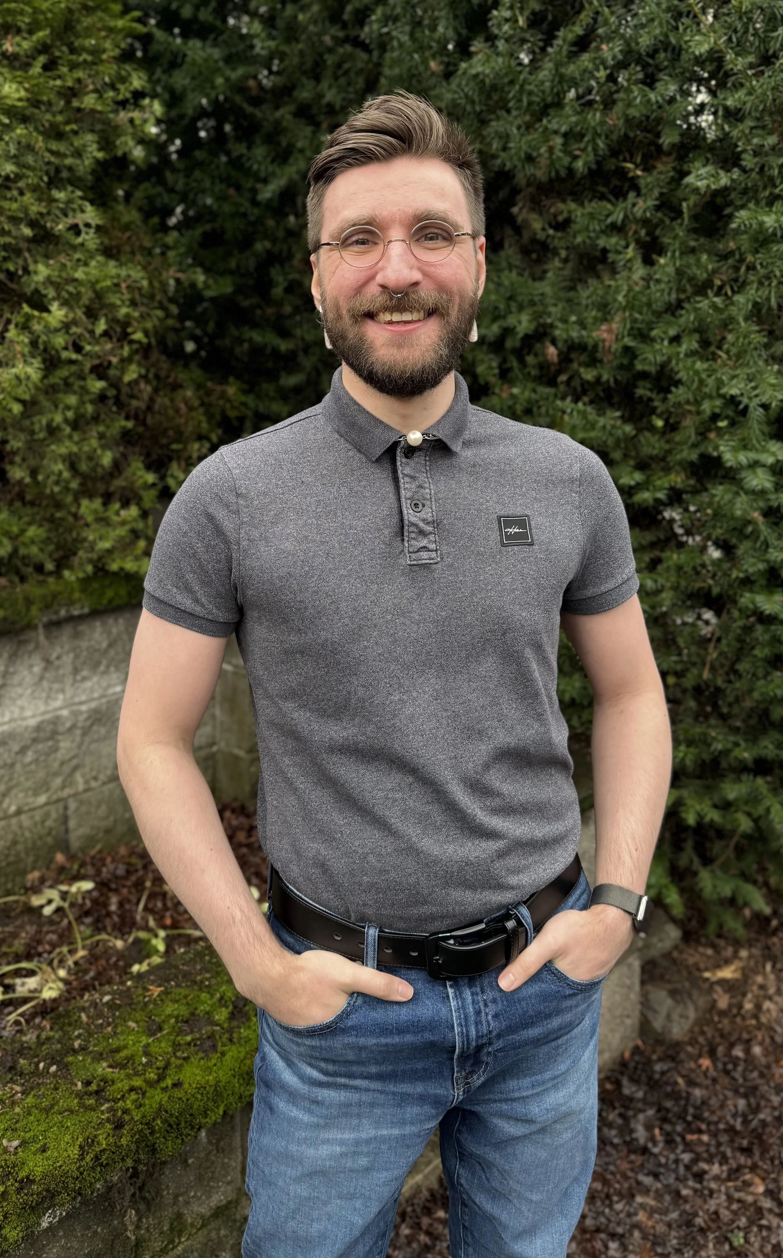 A smiling man with glasses and a beard stands outdoors in front of green bushes, wearing a gray polo shirt and blue jeans.