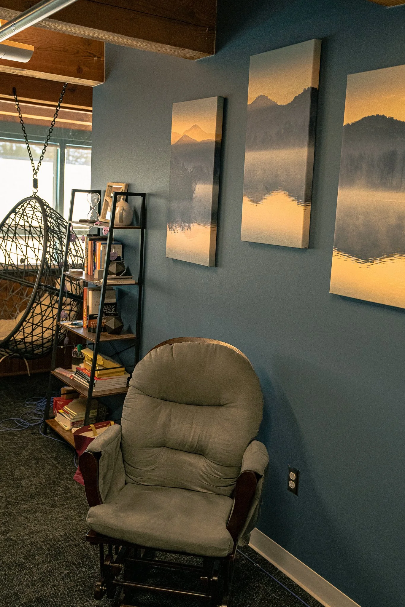 A cozy living room corner with a cushioned armchair, a black bookshelf with books and decorative items, a hanging egg chair, and three landscape canvas prints on a blue wall.