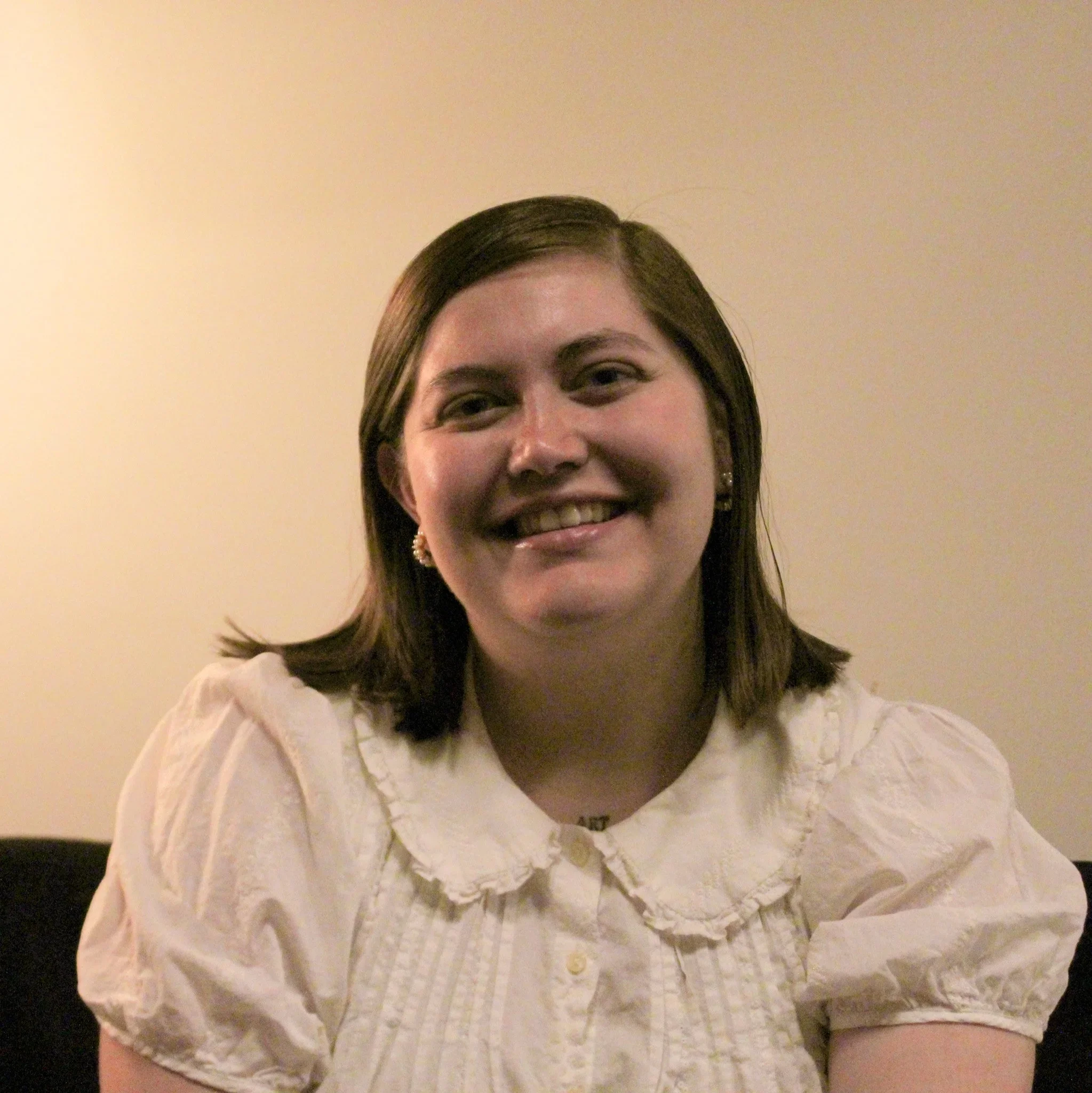 A woman with shoulder-length brown hair smiling, wearing a white blouse and earrings, sitting against a plain wall.