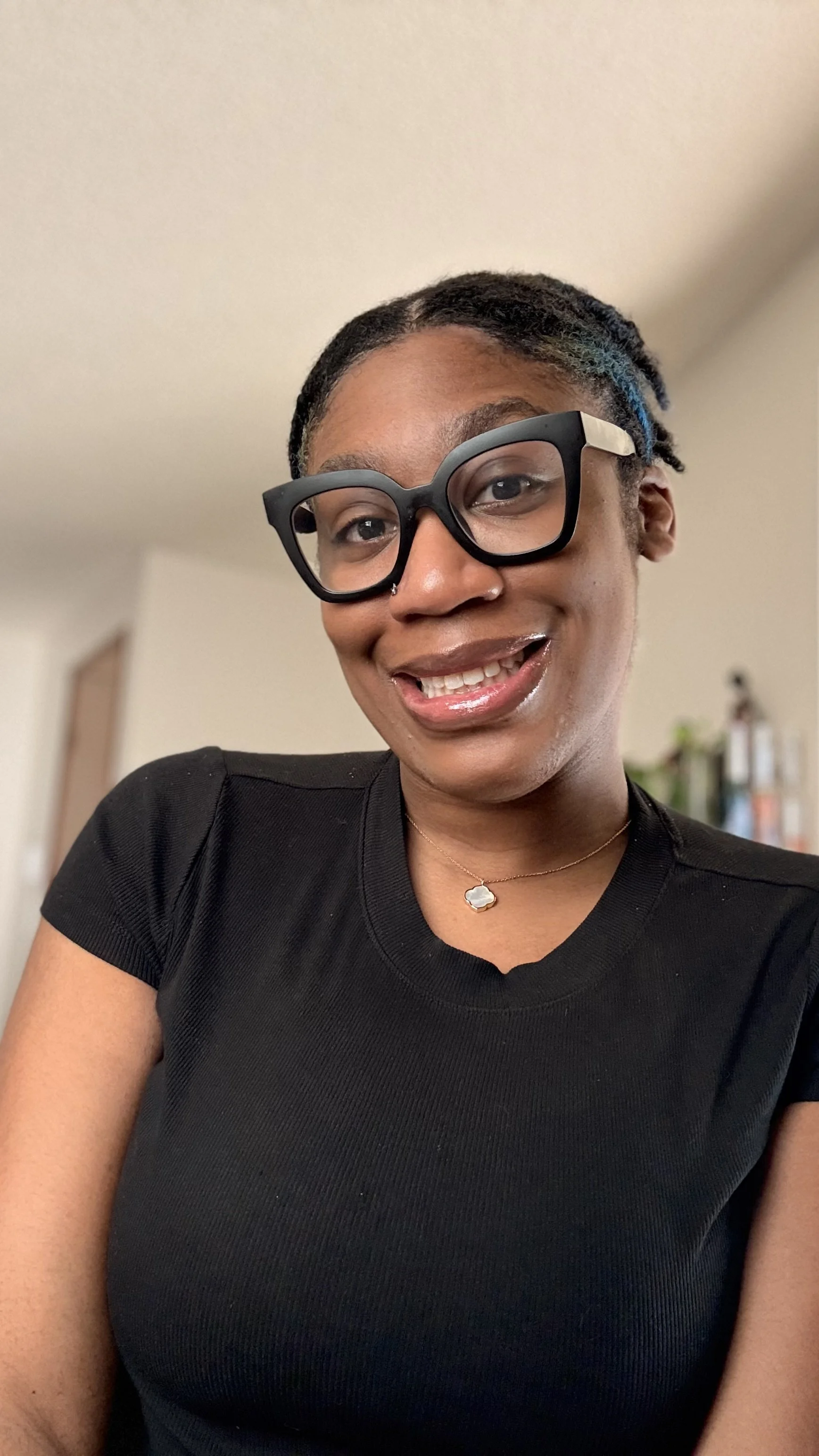 A smiling woman with glasses and a black shirt, wearing a necklace, in a home setting.
