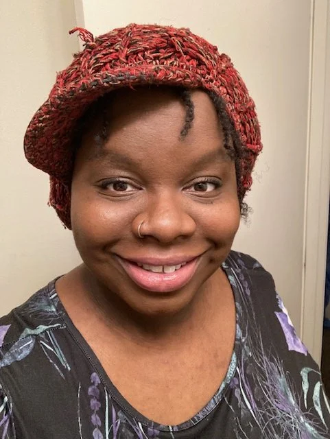 A woman with short curly hair wearing a colorful head wrap and a floral top, smiling at the camera.
