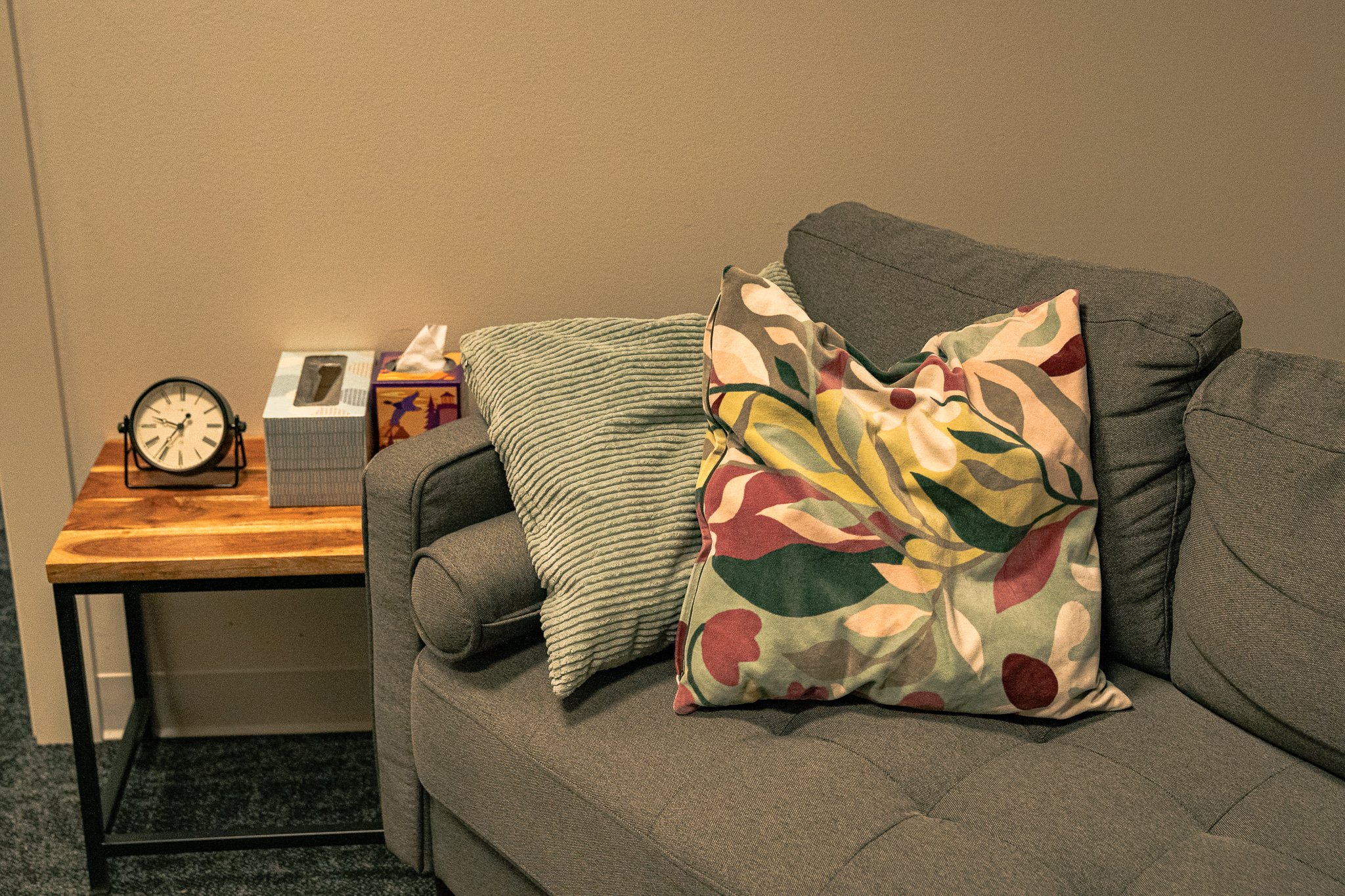 A gray sofa with two decorative pillows, one striped and one with a colorful leaf pattern, next to a wooden side table with a clock, tissue box, and a decorative item.