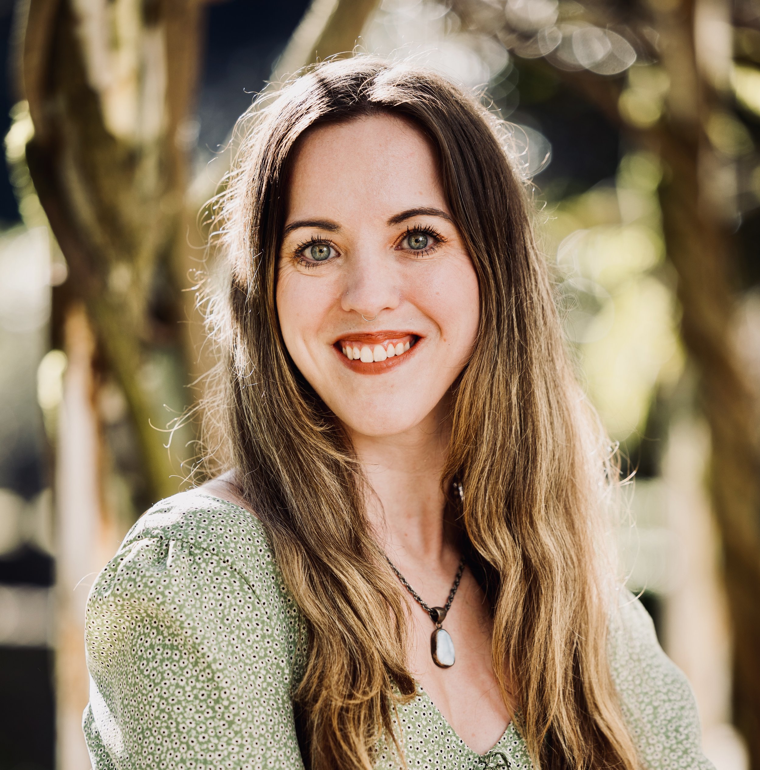 A young woman with long brown hair, green eyes, and a septum piercing smiling outdoors with trees in the background.