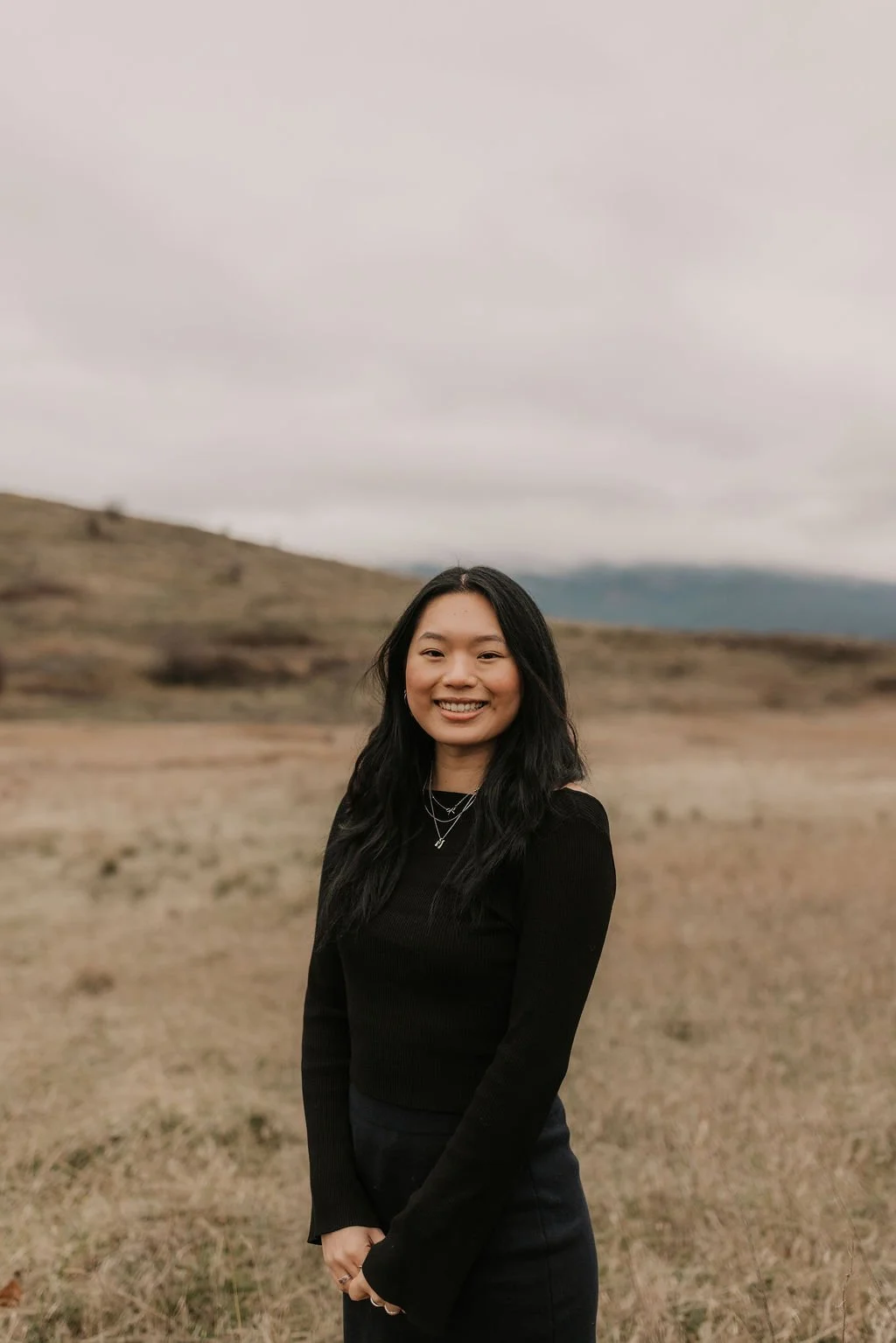 A woman with long black hair smiling outdoors in a vast, open landscape with hills and cloudy sky.