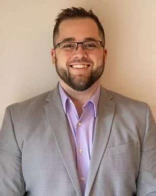 Portrait of a man with glasses and a beard, wearing a light gray suit and a purple shirt, smiling against a plain background.