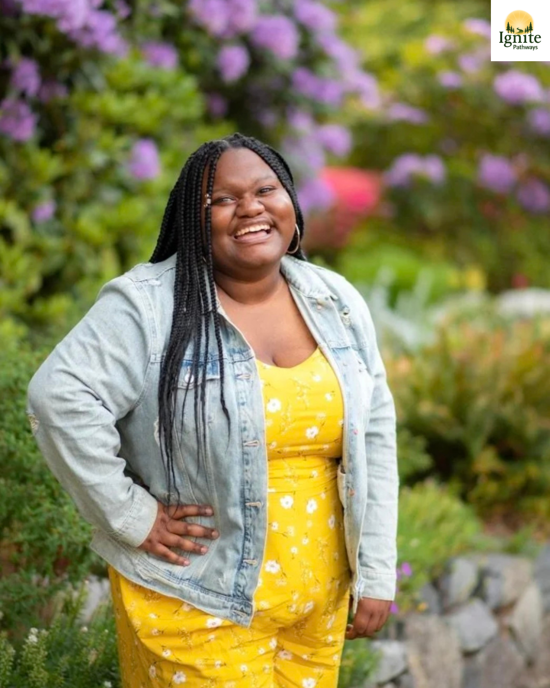 Woman with long braided hair, wearing a yellow floral dress and denim jacket, smiling outdoors with purple flowers in the background.