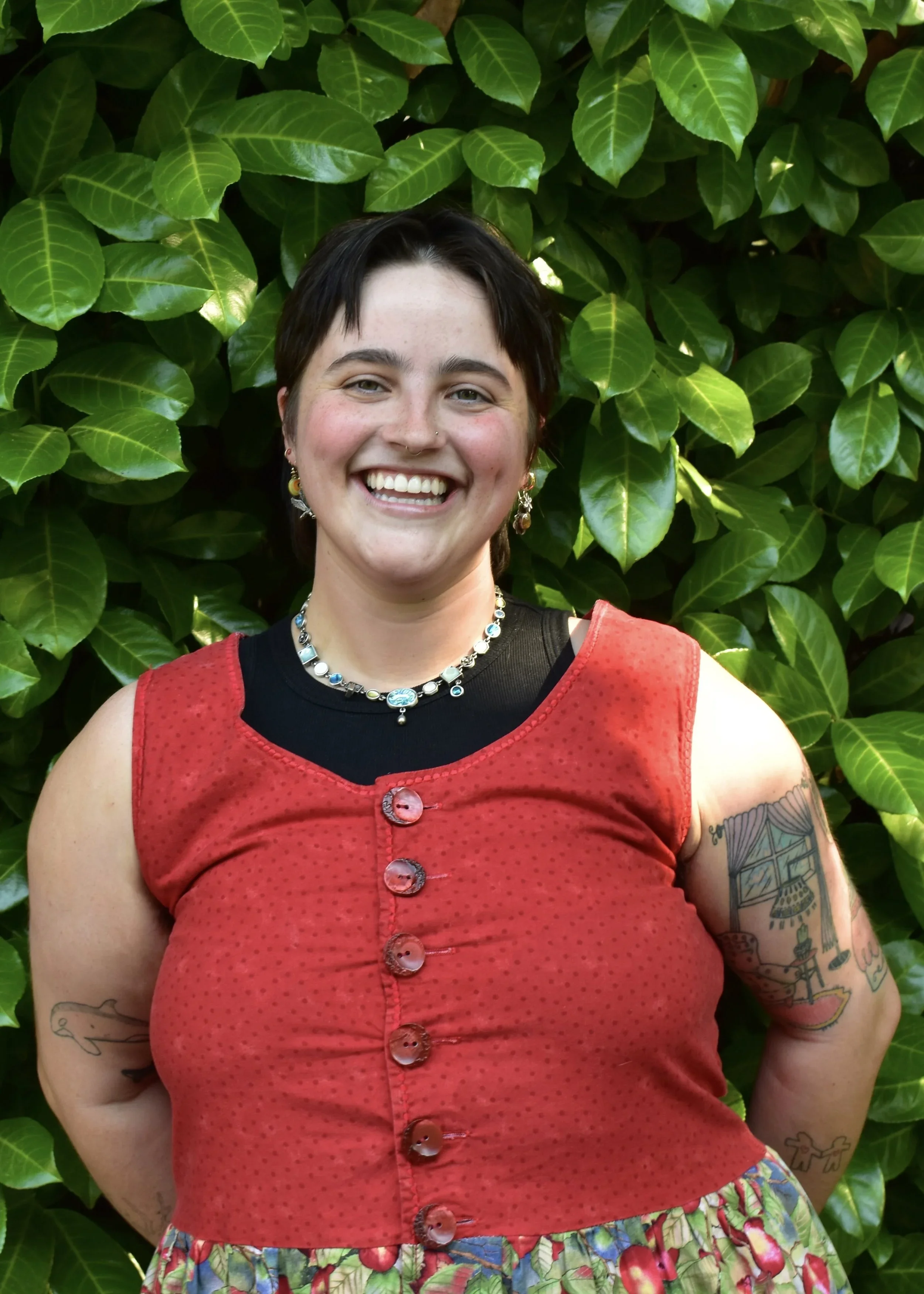 A smiling woman with short dark hair, tattoos on her arms, wearing a red sleeveless top with buttons, a black shirt underneath, a colorful skirt, and jewelry, standing in front of green leafy plants.