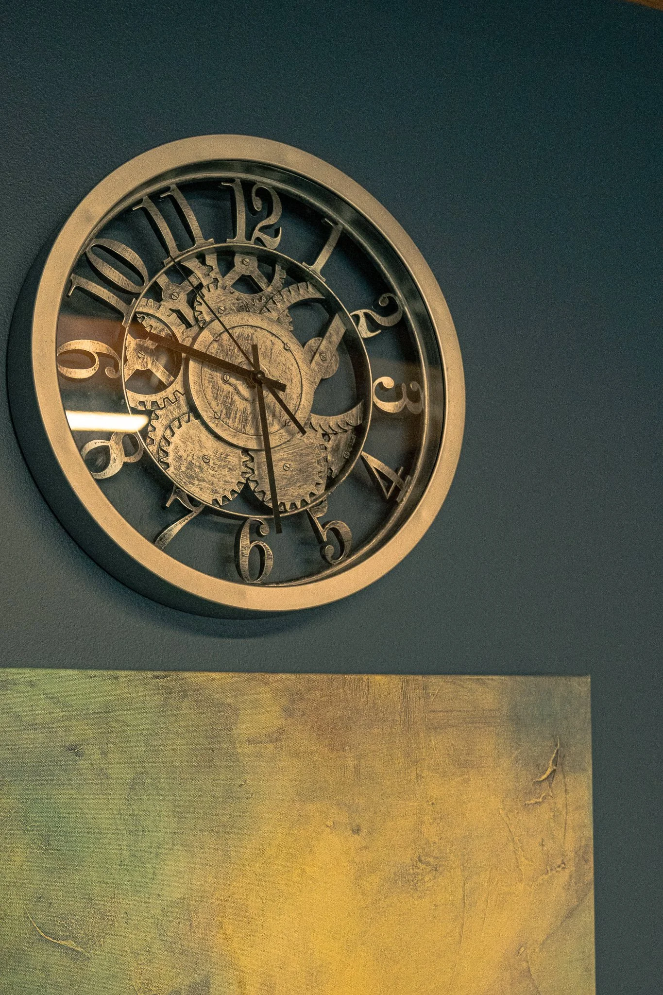 A round wall clock with visible gears, mounted on a dark blue wall above a yellowish-green textured surface.