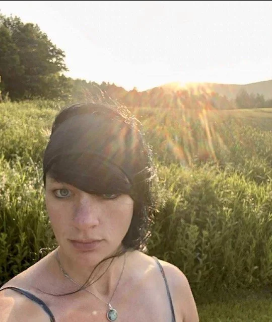 Young woman with black hair and blue eyes taking a selfie outdoors during sunset in a grassy field with trees in the background.
