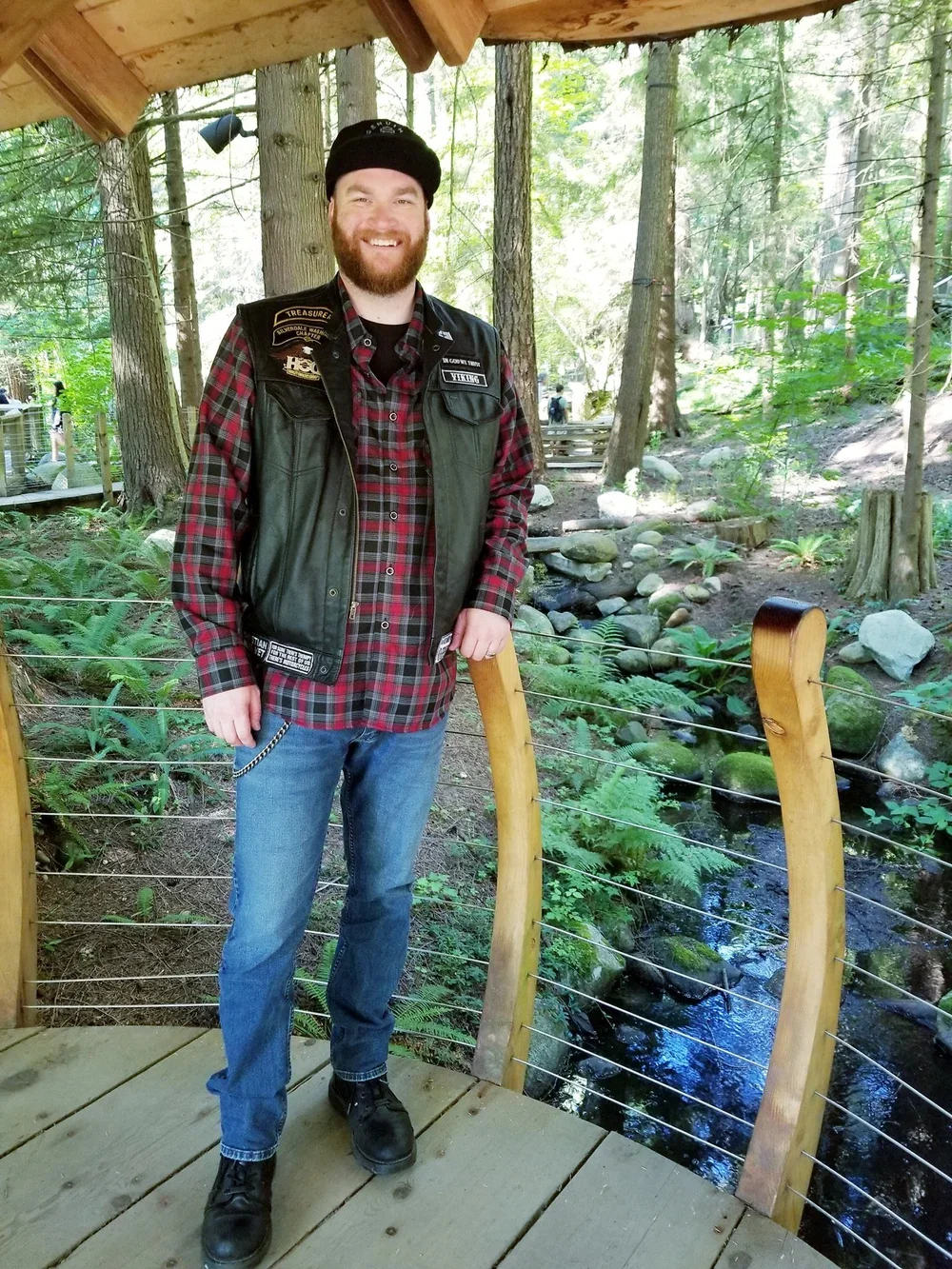 Smiling man with a beard in a forested area, wearing a black beanie, a black vest with patches, a red and black plaid shirt, blue jeans, and black boots, standing on a wooden platform near a small stream.