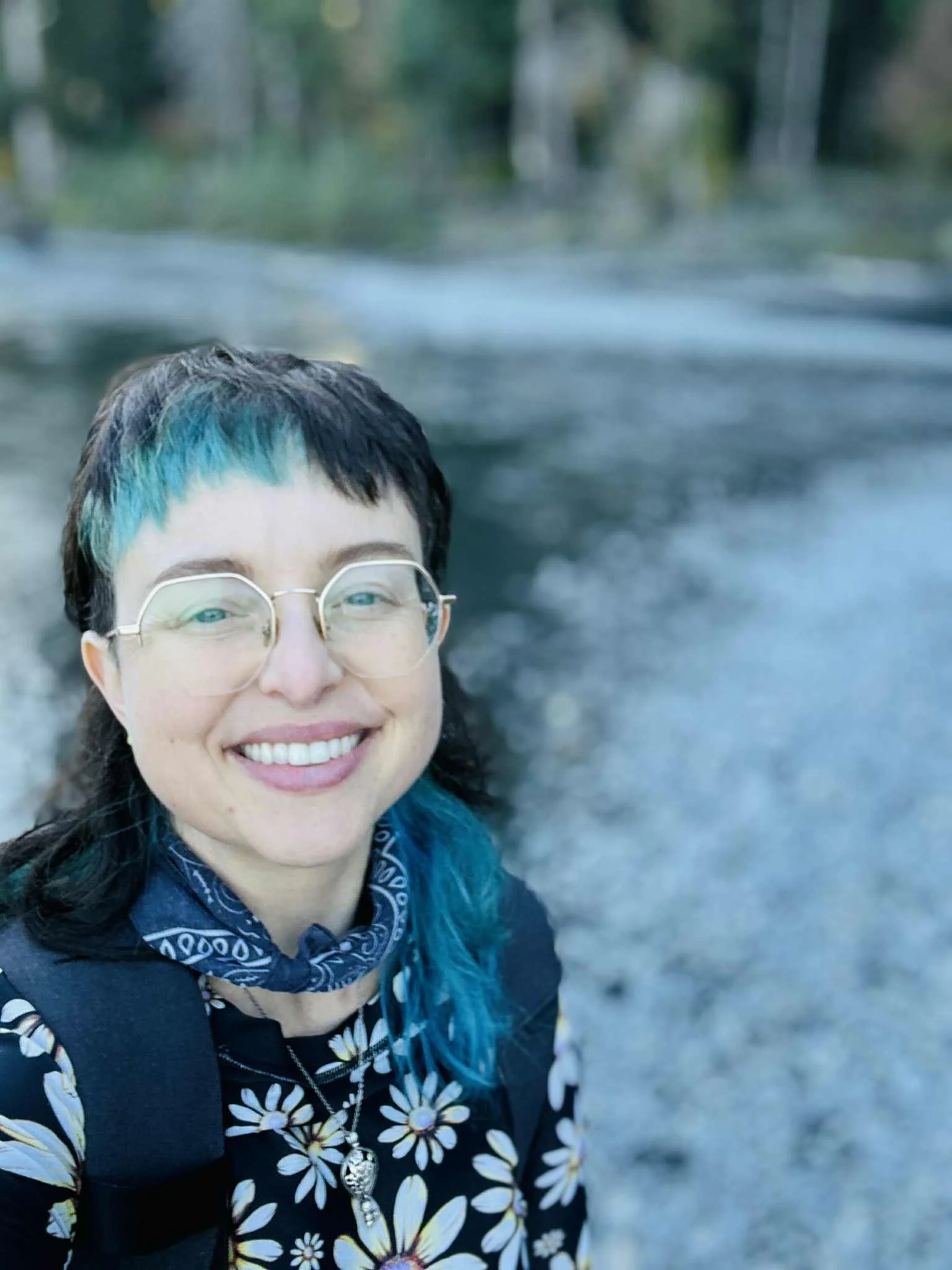 A woman smiling while taking a selfie outdoors near a river or stream.