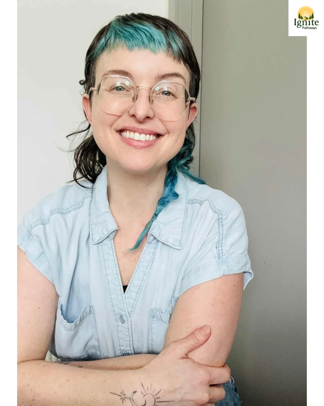 A smiling woman with short dark hair and turquoise highlights, wearing glasses and a light denim sleeveless shirt, sitting with arms crossed, showing a moon and star tattoo on her arm, in front of a plain background.
