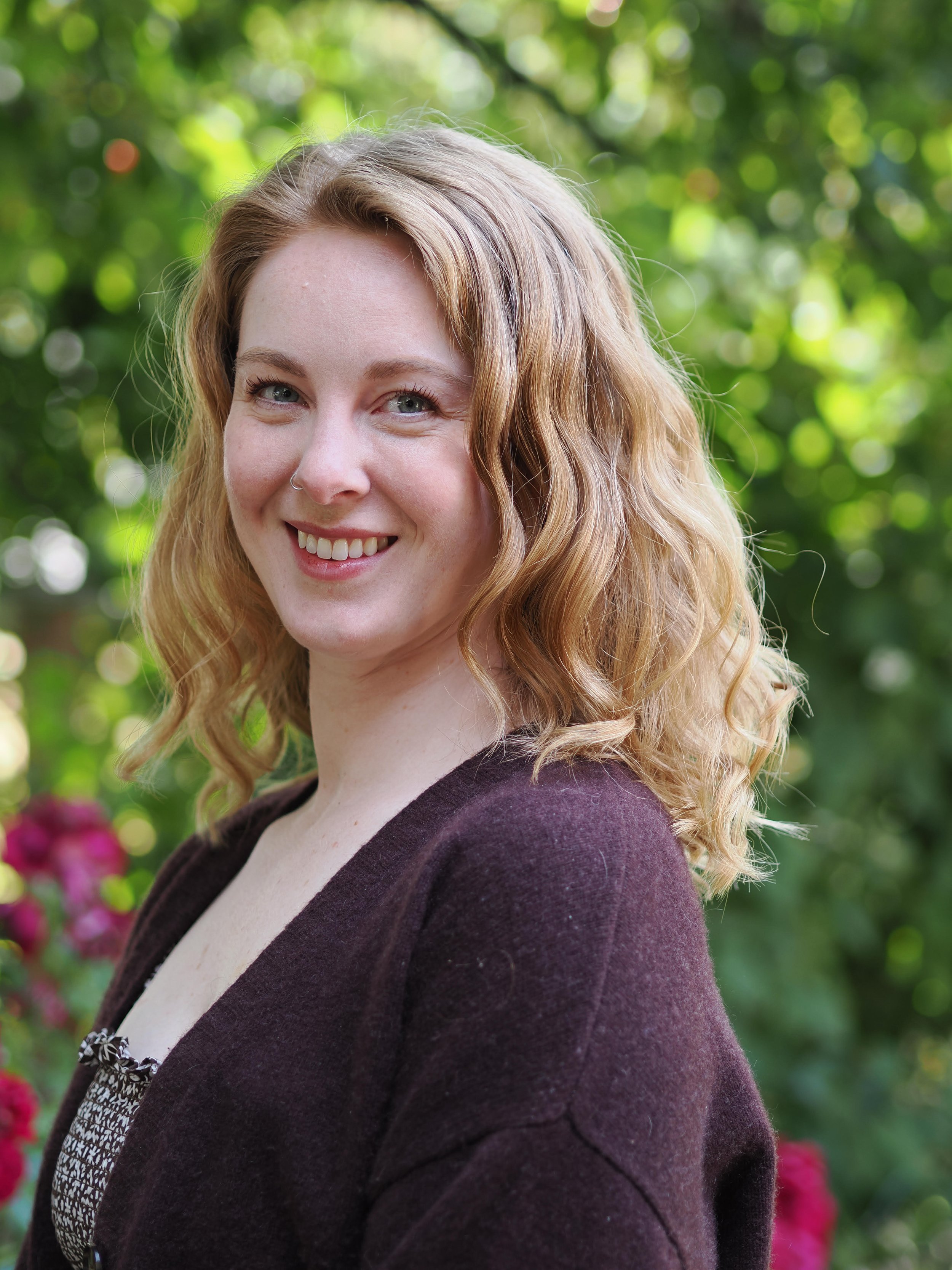 A portrait of a young woman with wavy blonde hair, smiling, outdoors with green foliage in the background.
