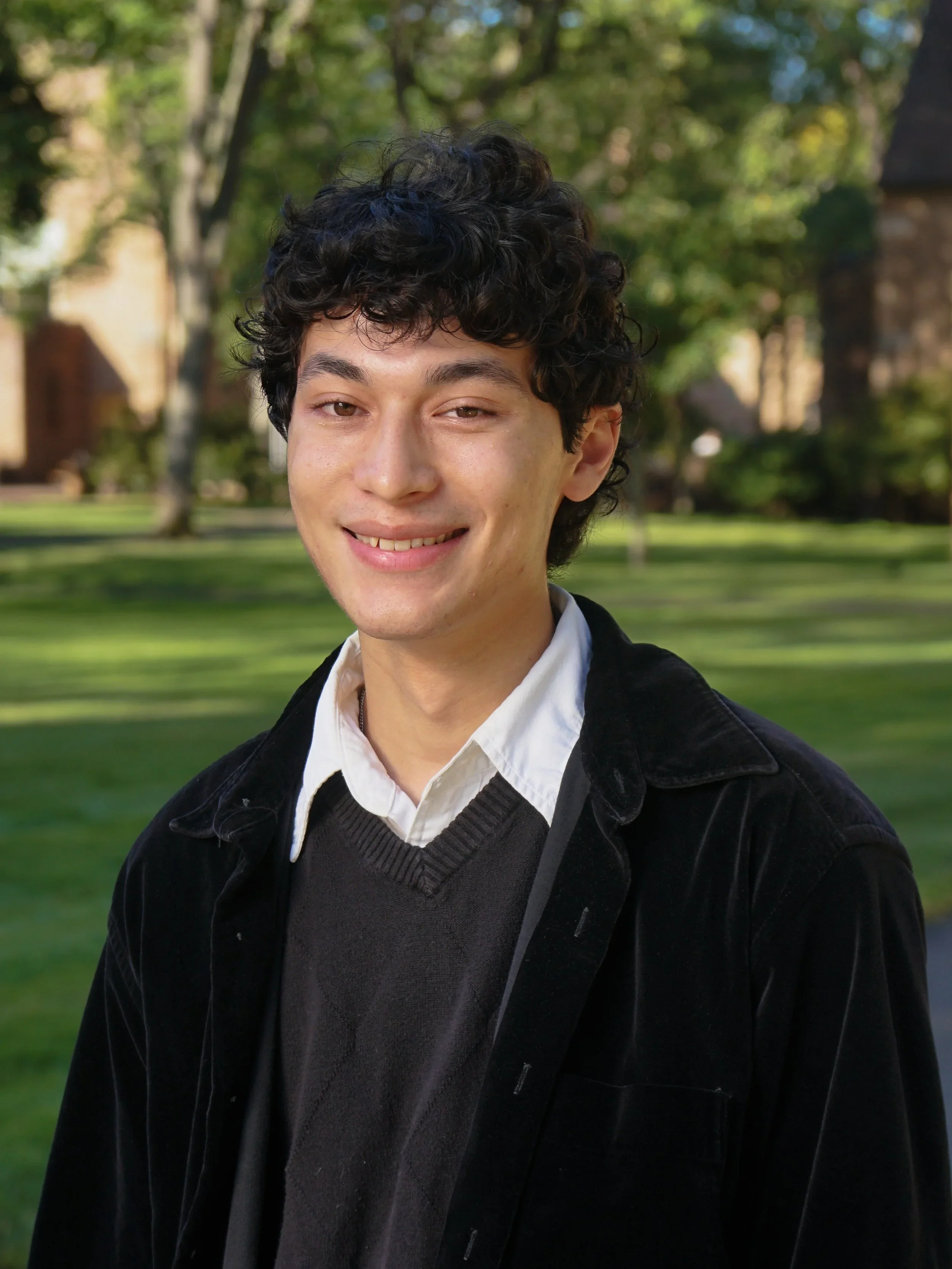 A young man with curly black hair, wearing a white shirt and a dark sweater, smiling outdoors with trees and grass in the background.