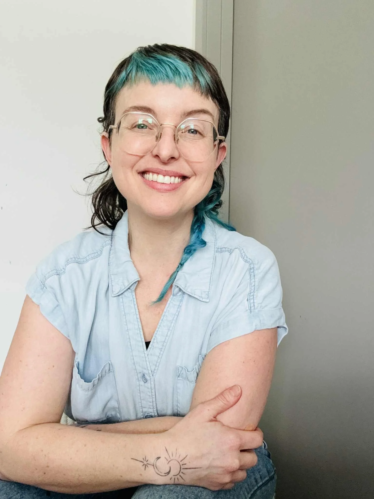 A woman with short hair dyed turquoise and dark brown, wearing glasses and a light denim shirt, sitting with her arms crossed and smiling.
