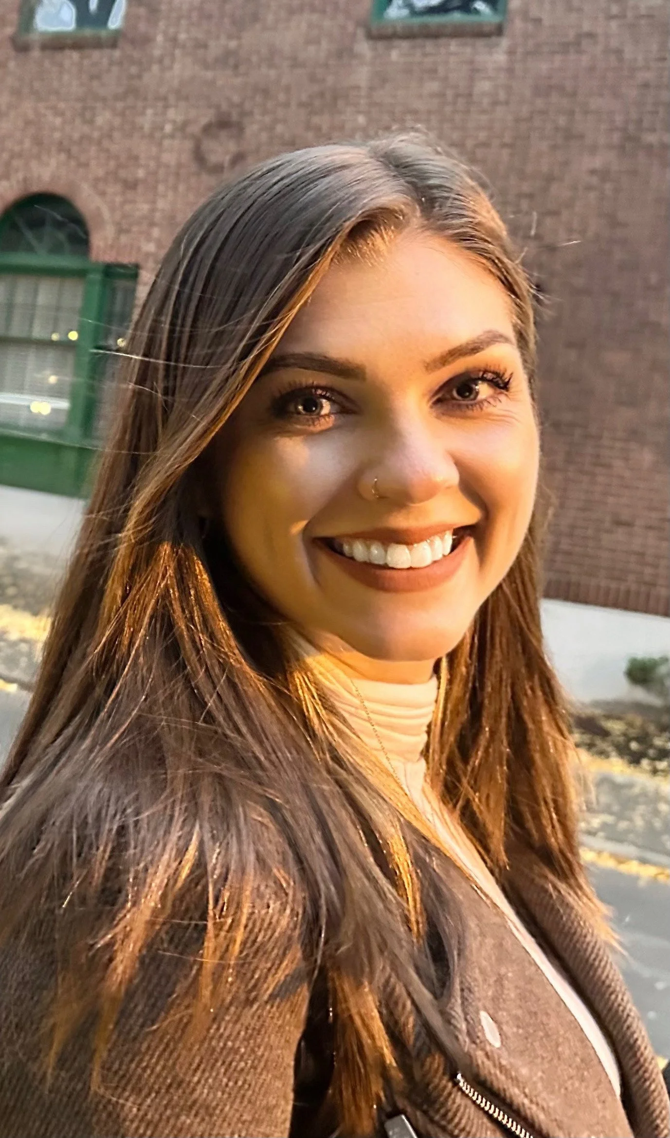 Close-up of a smiling woman with long brown hair, wearing a brown jacket, standing outside at dusk with a brick building in the background.