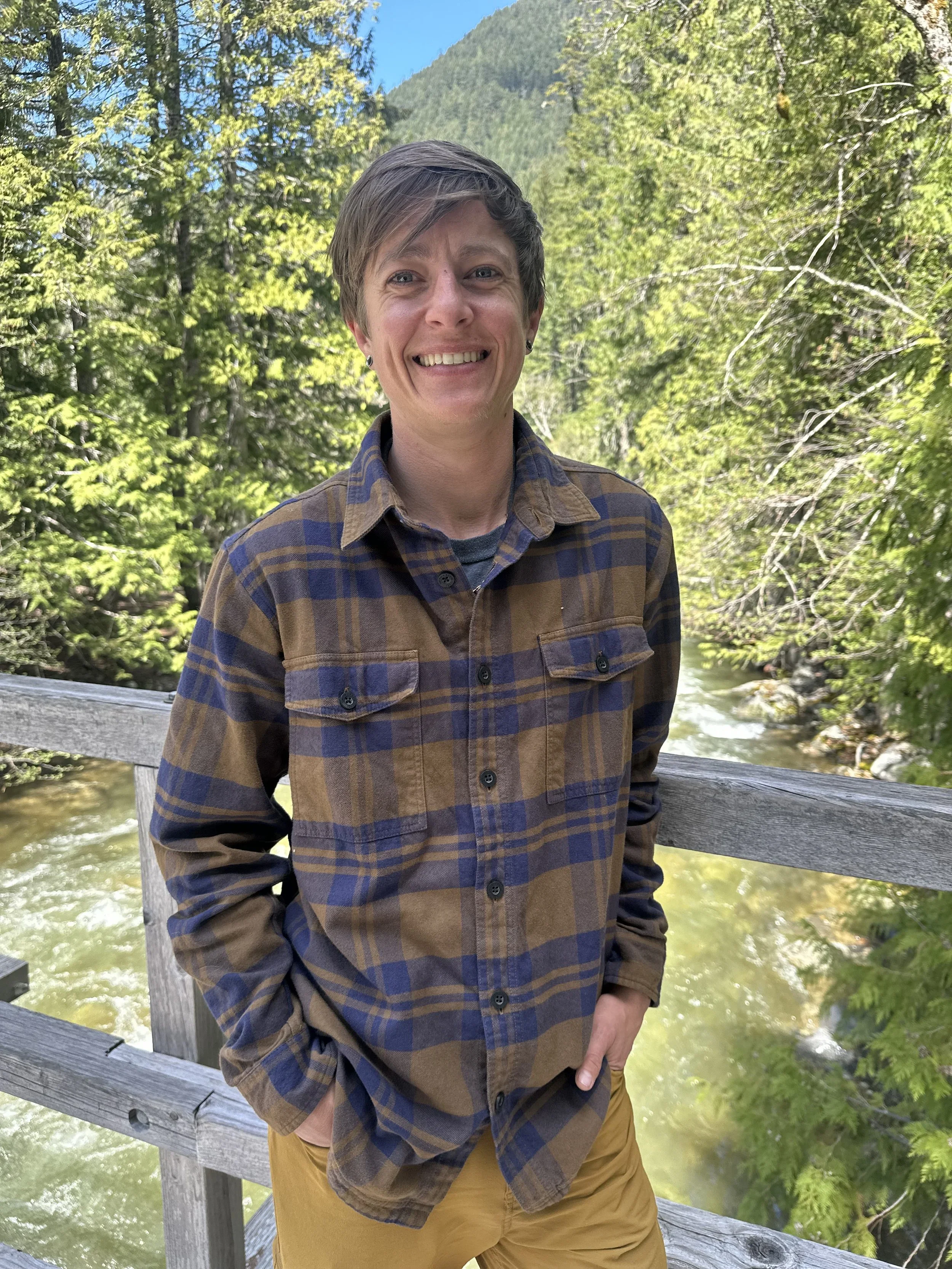 A person with short brown hair, wearing a checkered brown and blue shirt and mustard yellow pants, standing outdoors on a wooden bridge with a mountain and lush green trees in the background.