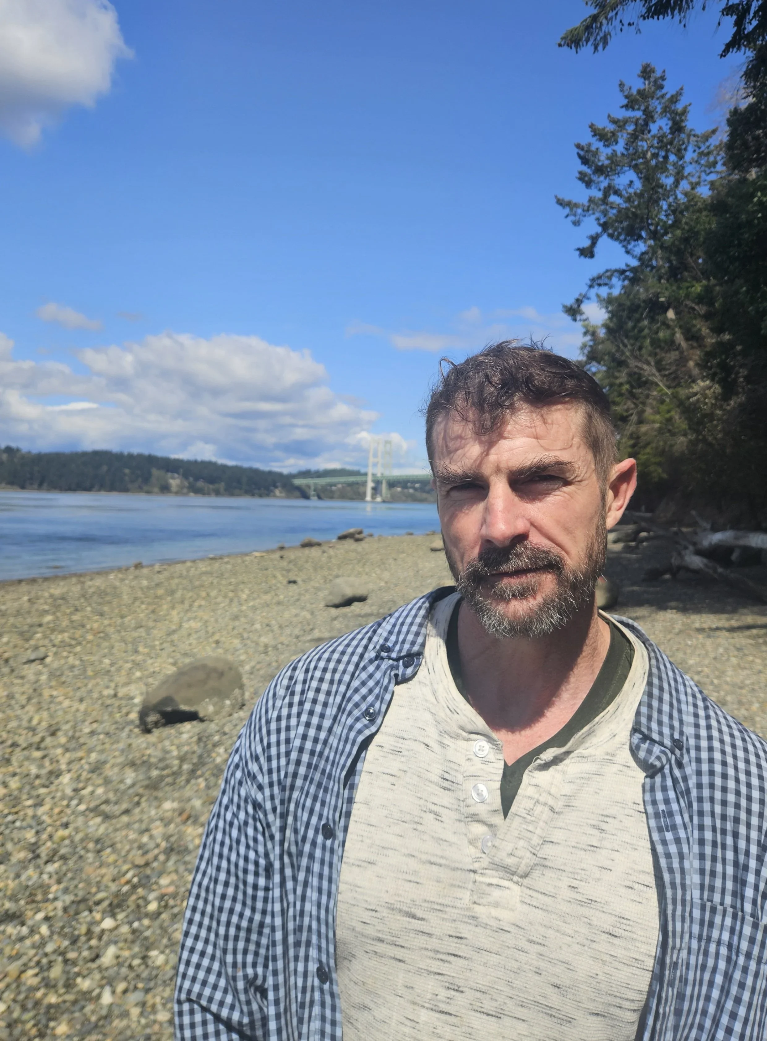 A man with a beard and short hair standing on a rocky shoreline near a body of water, with a bridge in the background and trees to the right, during a sunny day with blue sky and some clouds.
