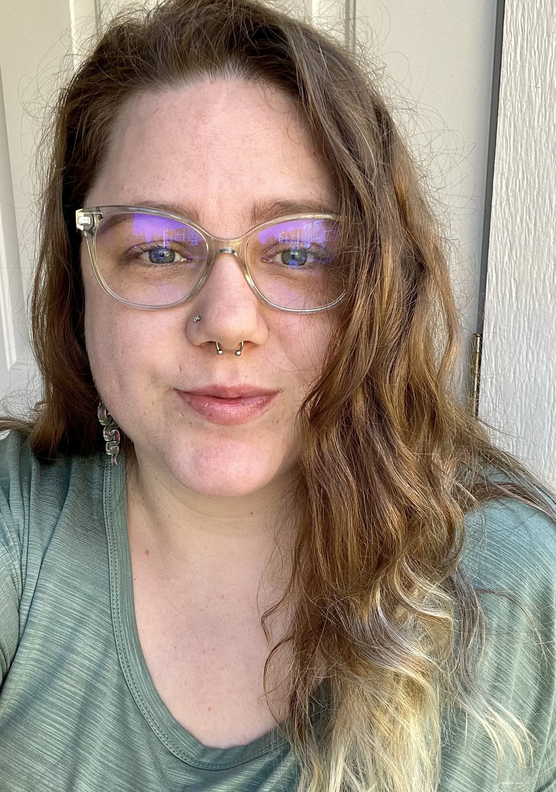 A woman with wavy, reddish-brown hair, wearing glasses, a nose piercing, and earrings, standing near a window with natural light.