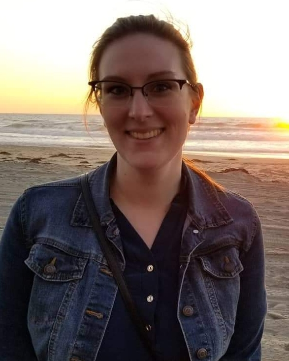 A smiling woman wearing glasses and a denim jacket standing on a beach at sunset, with the ocean and sky in the background.