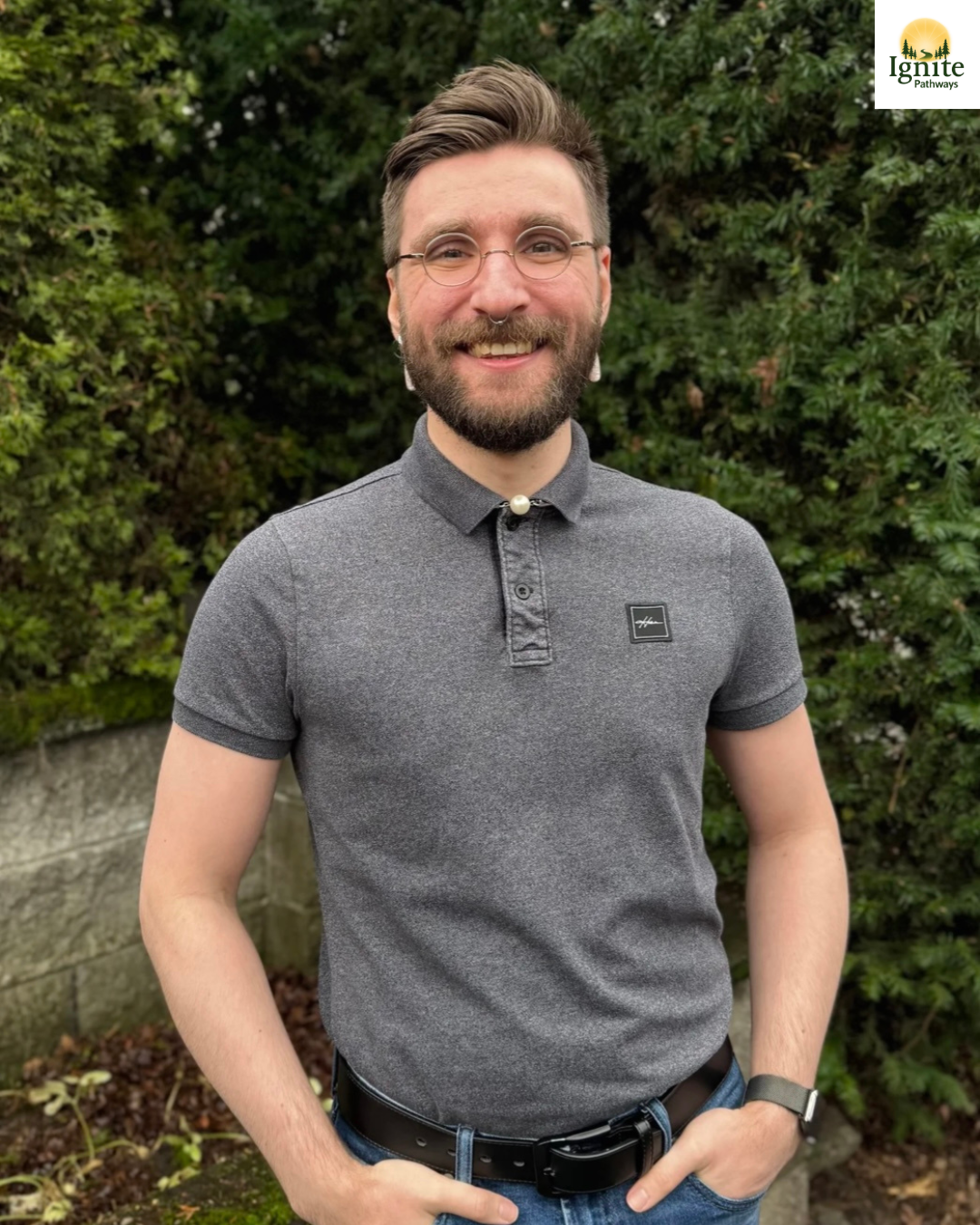 A smiling man with glasses and a beard standing outdoors in front of green bushes. He is wearing a gray polo shirt with a small logo, a belt, and a watch, and has one hand in his pocket.