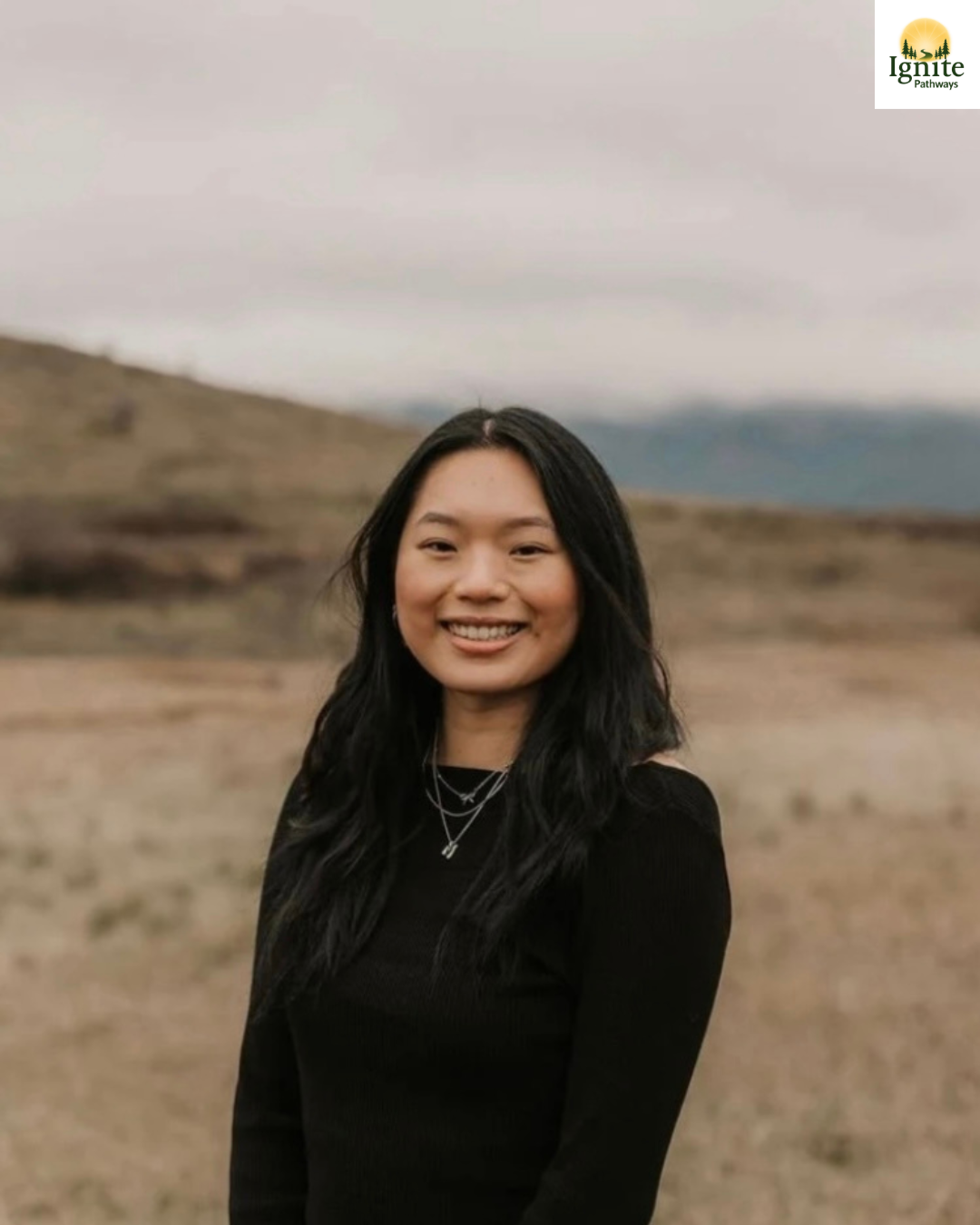 A woman smiling outdoors on a cloudy day with rolling hills and a cloudy sky in the background.