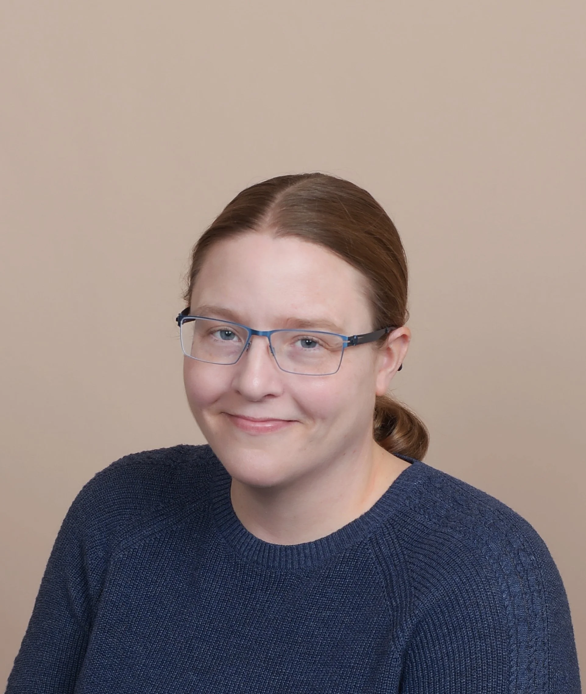 A young woman with red hair, glasses, and a blue sweater smiling at the camera against a beige background.
