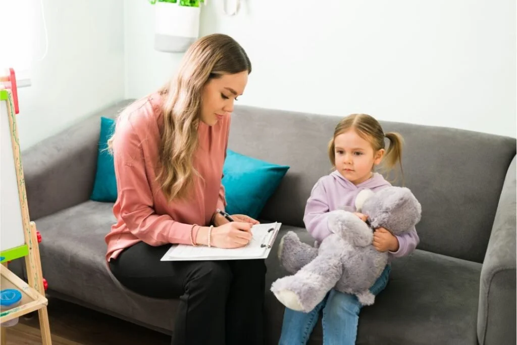 A woman sitting on a gray couch with a clipboard, talking to a young girl with pigtails holding a teddy bear in a cozy room.