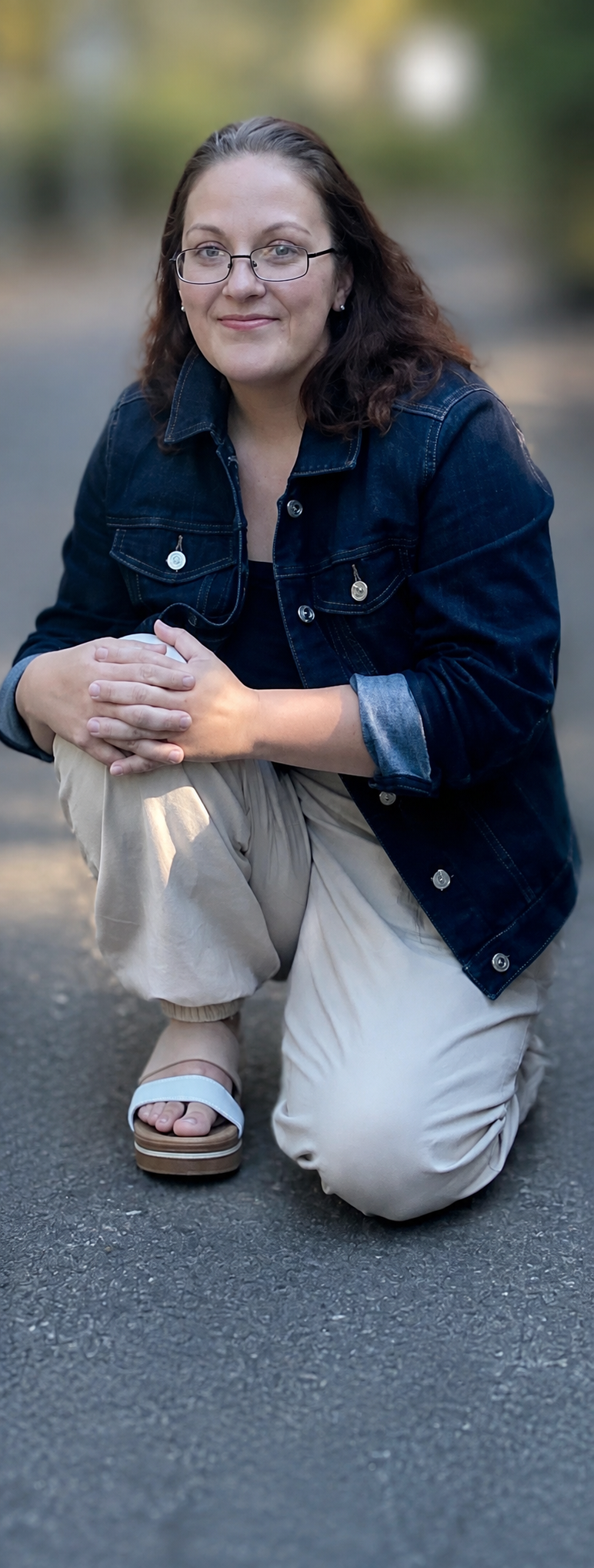 A woman with glasses, wearing a denim jacket and beige pants is kneeling on the ground outdoors, smiling at the camera.