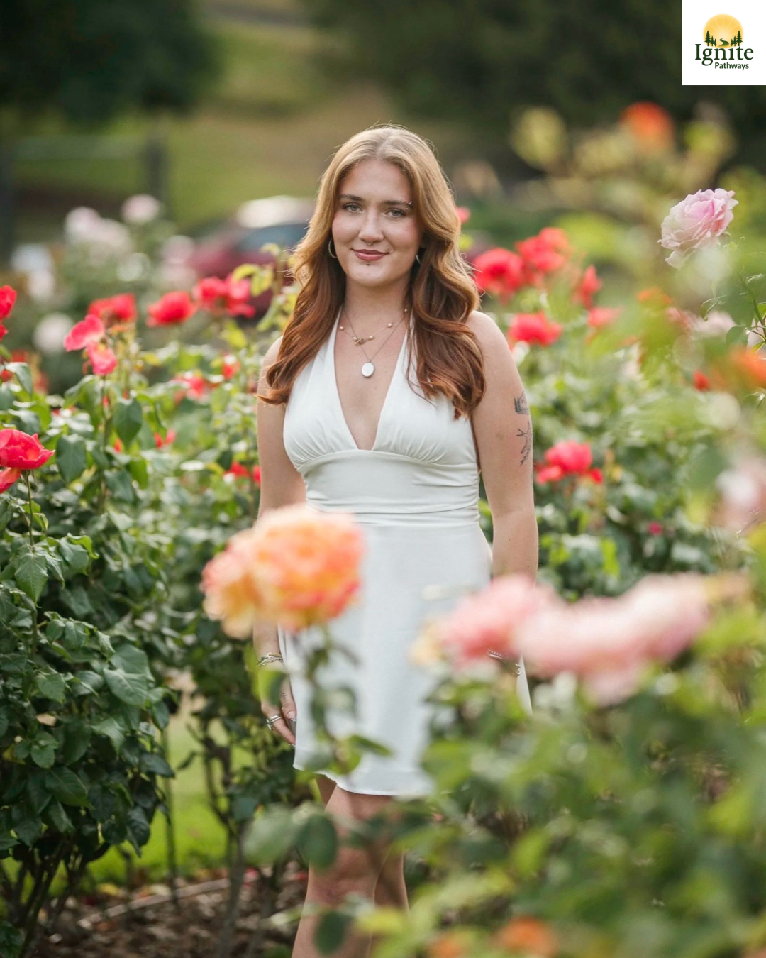 A woman standing among blooming roses in a garden, wearing a white dress and jewelry, smiling at the camera.
