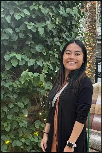 A smiling woman with long black hair standing next to a leafy green plant with small lights, outdoors in a city environment.