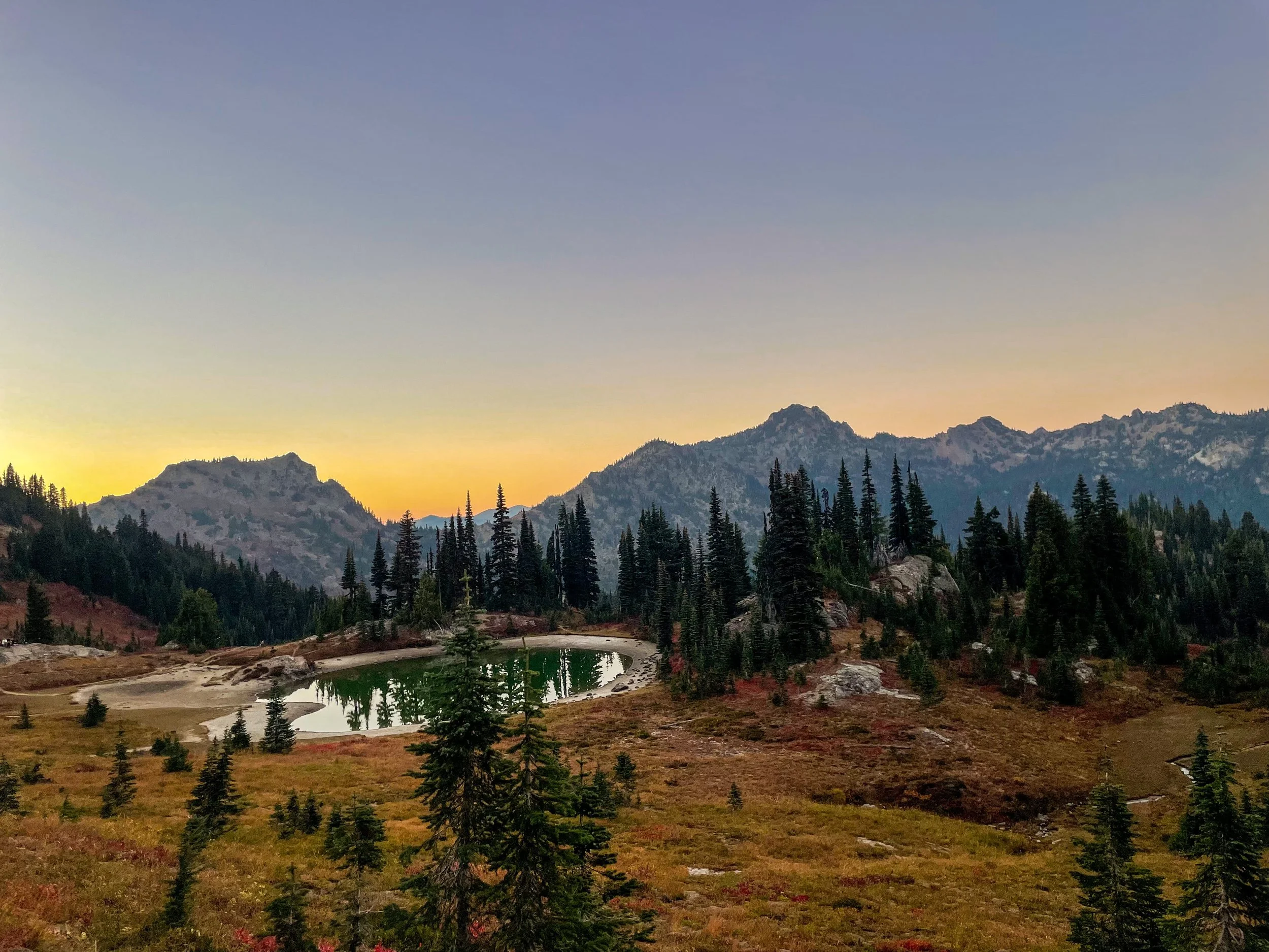 A scenic mountain landscape at sunset with a small pond, pine trees, and rugged mountains in the distance.