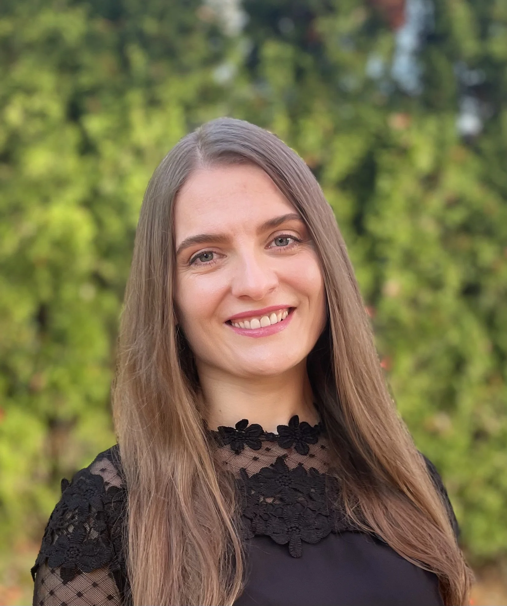 Portrait of a young woman with long brown hair, smiling, wearing a black lace top, outdoors with greenery in the background.