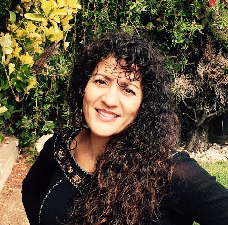 Yvette Fama, LMFT, a woman with curly dark hair smiling outdoors surrounded by green foliage and plants. She offers emdr and somatic therapy in San Francisco, California and online via telehealth.