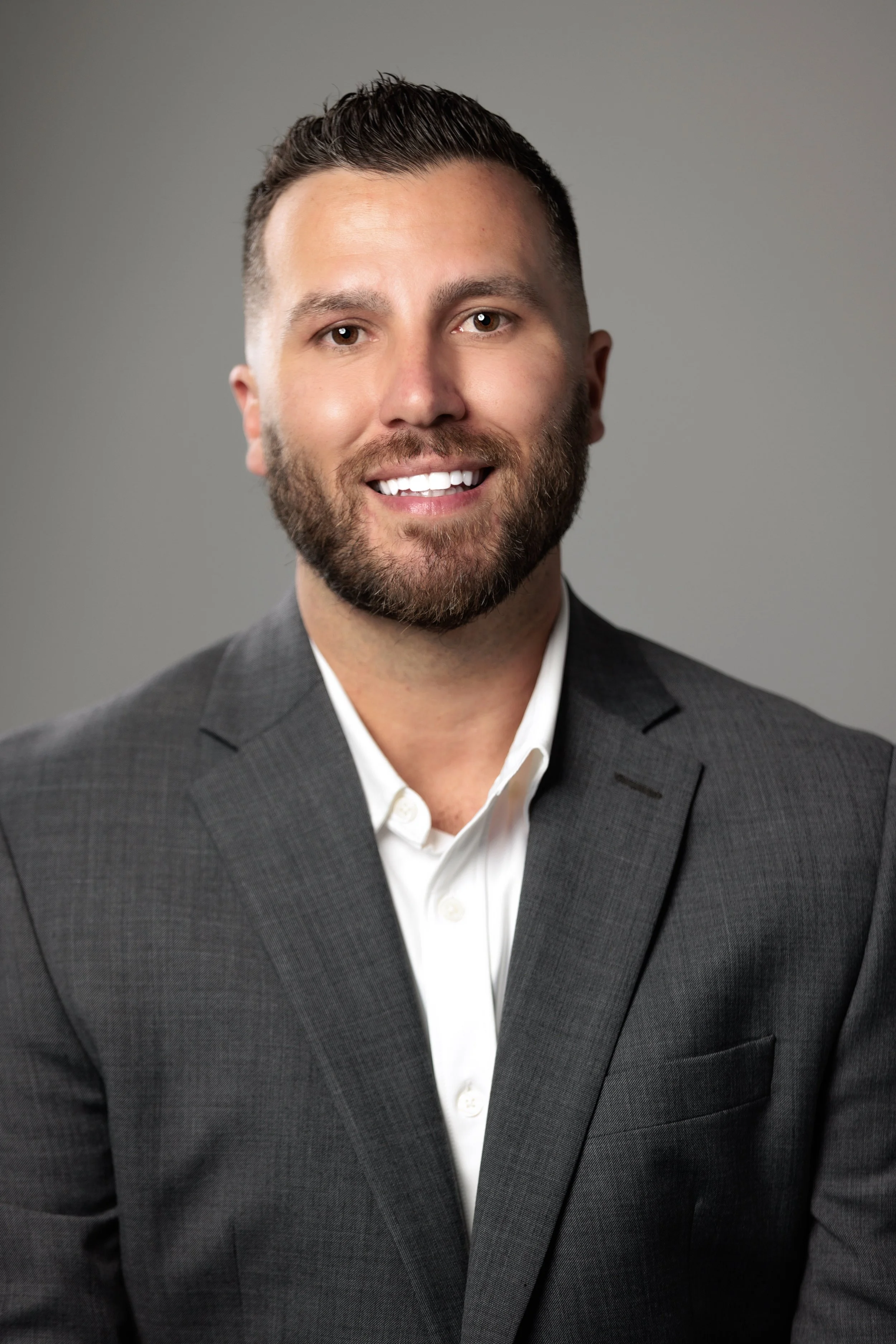 Headshot of a smiling man in a gray suit and white shirt, with short dark hair and a beard, against a gray background.