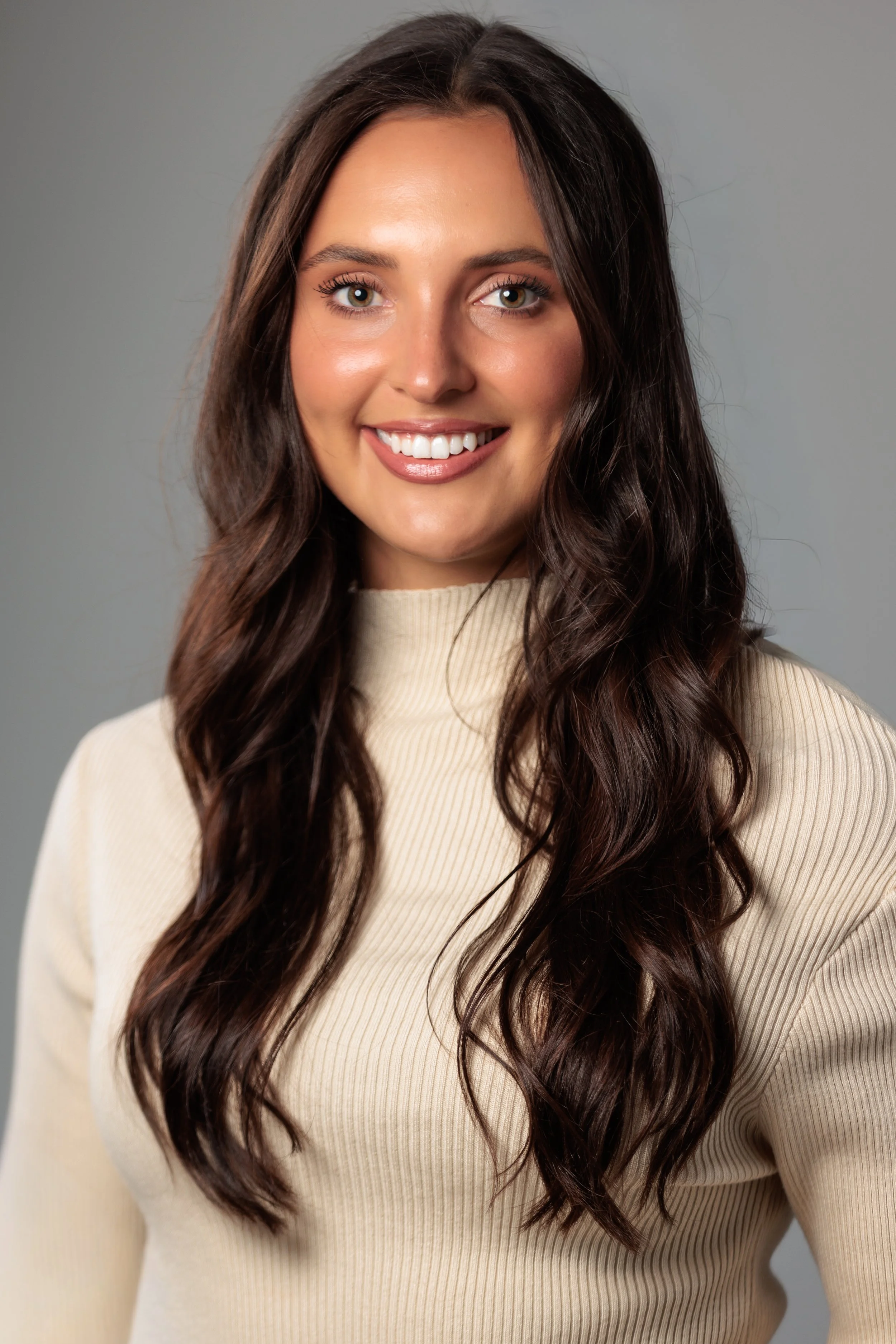 A young woman with long, wavy brown hair and light skin, smiling, wearing a beige turtleneck sweater, standing against a gray background.