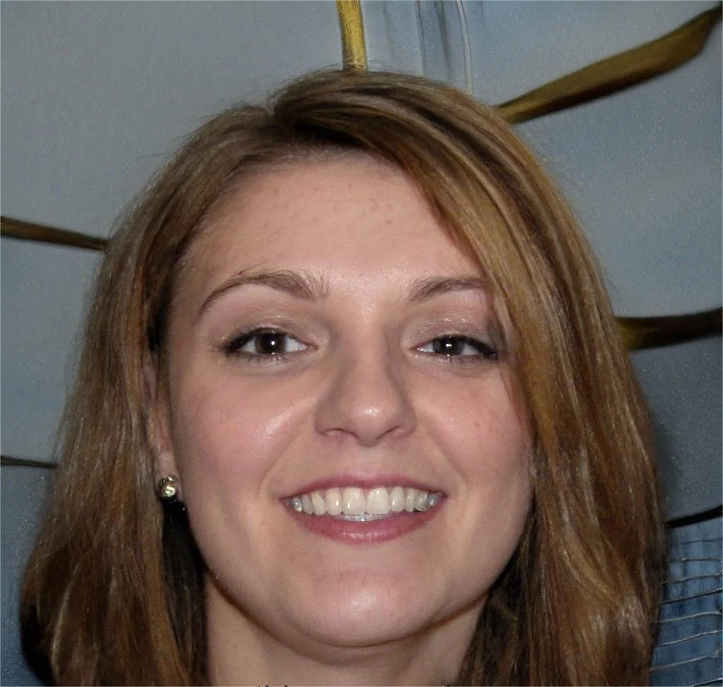 A woman with light brown hair, smiling, with a gray wall and wooden beams in the background.