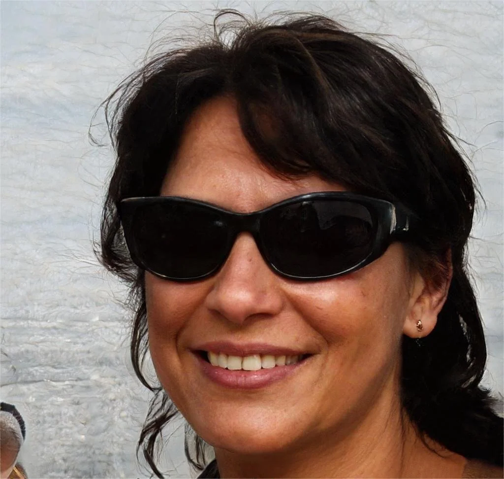 A woman with short dark hair wearing black sunglasses and smiling, with a light-colored background.
