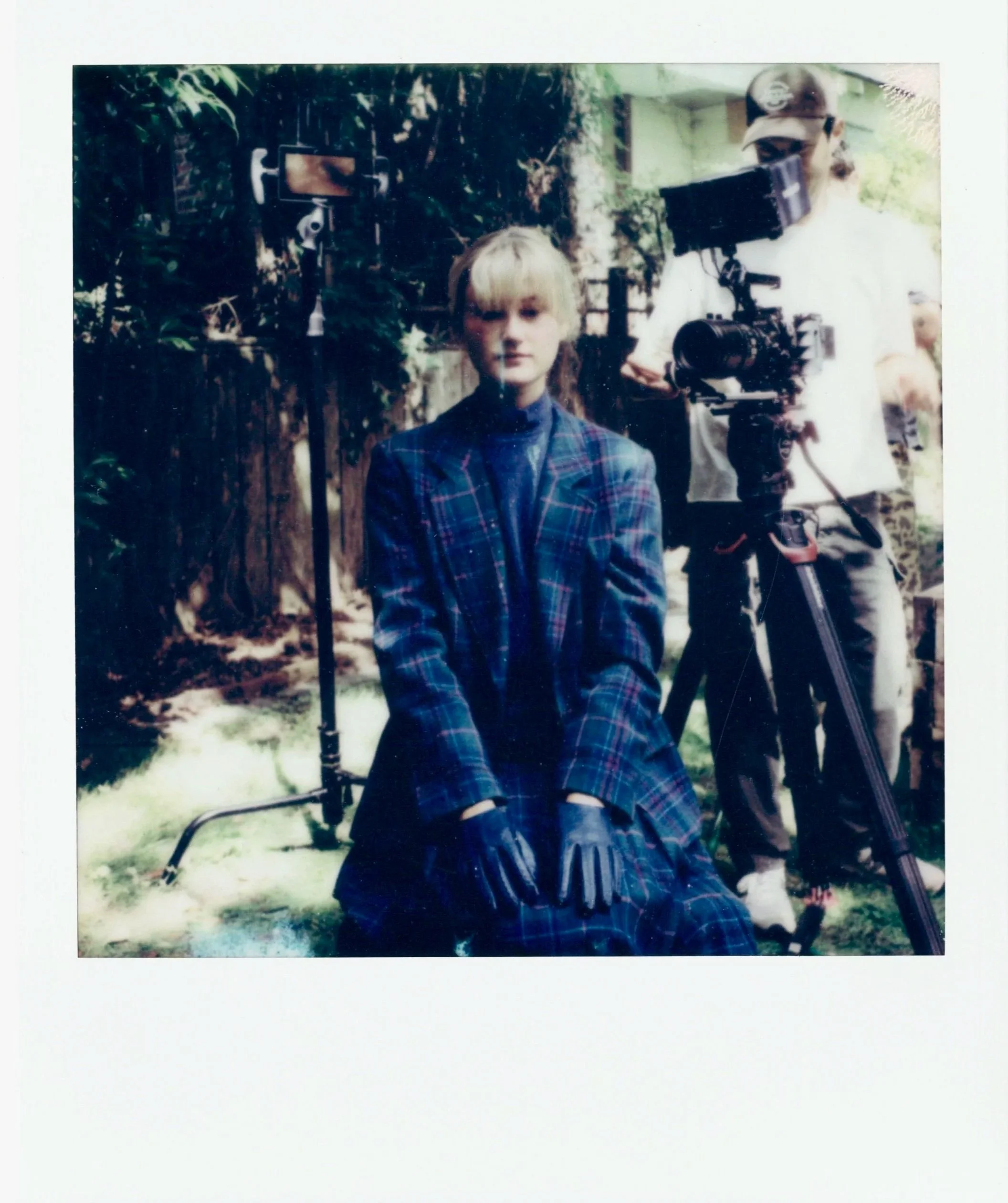 A young person dressed in a plaid suit and leather gloves is sitting in front of camera equipment, with a crew member operating the camera, outdoors in a garden.