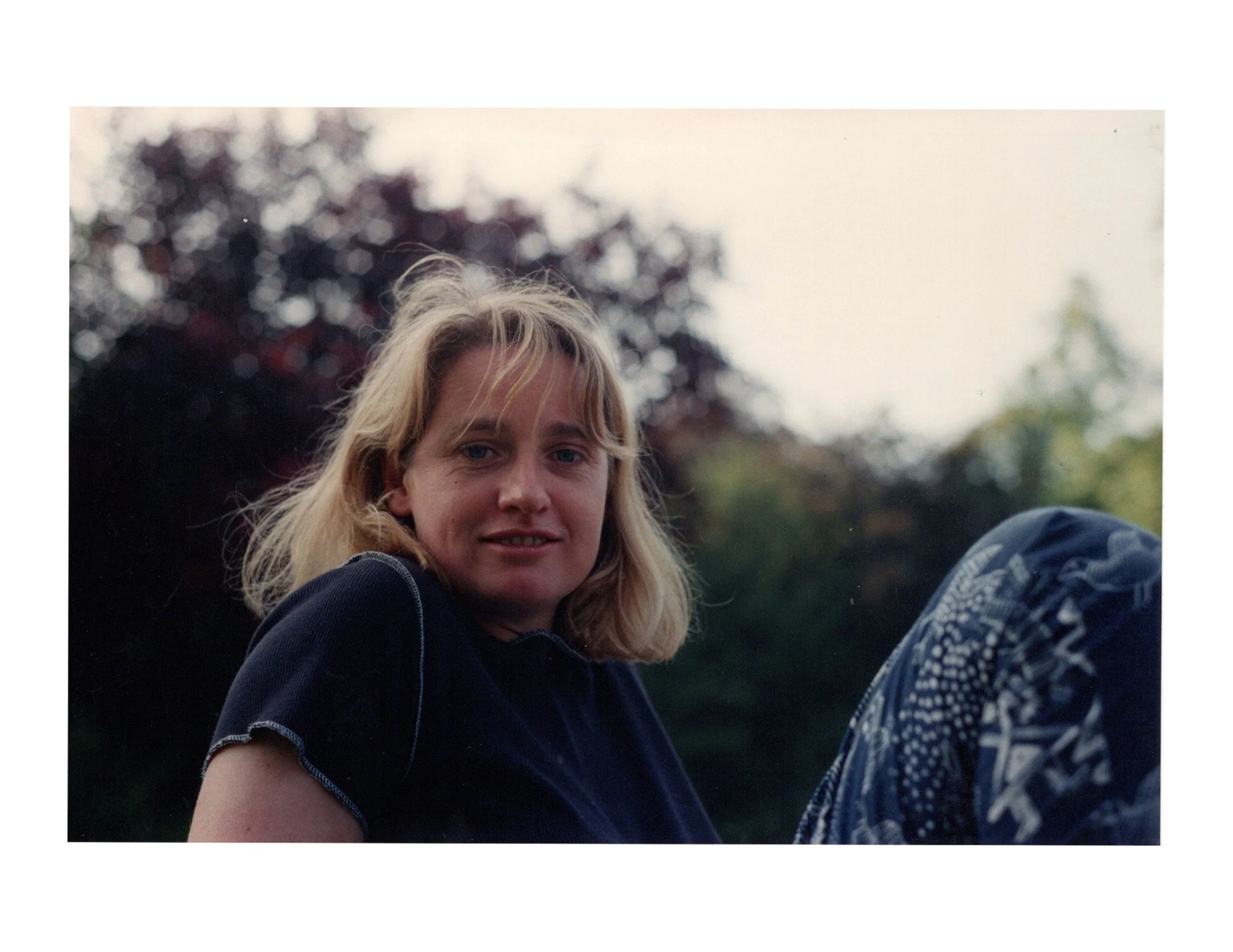 A woman with blonde hair, wearing a dark shirt, outdoors with trees in the background.