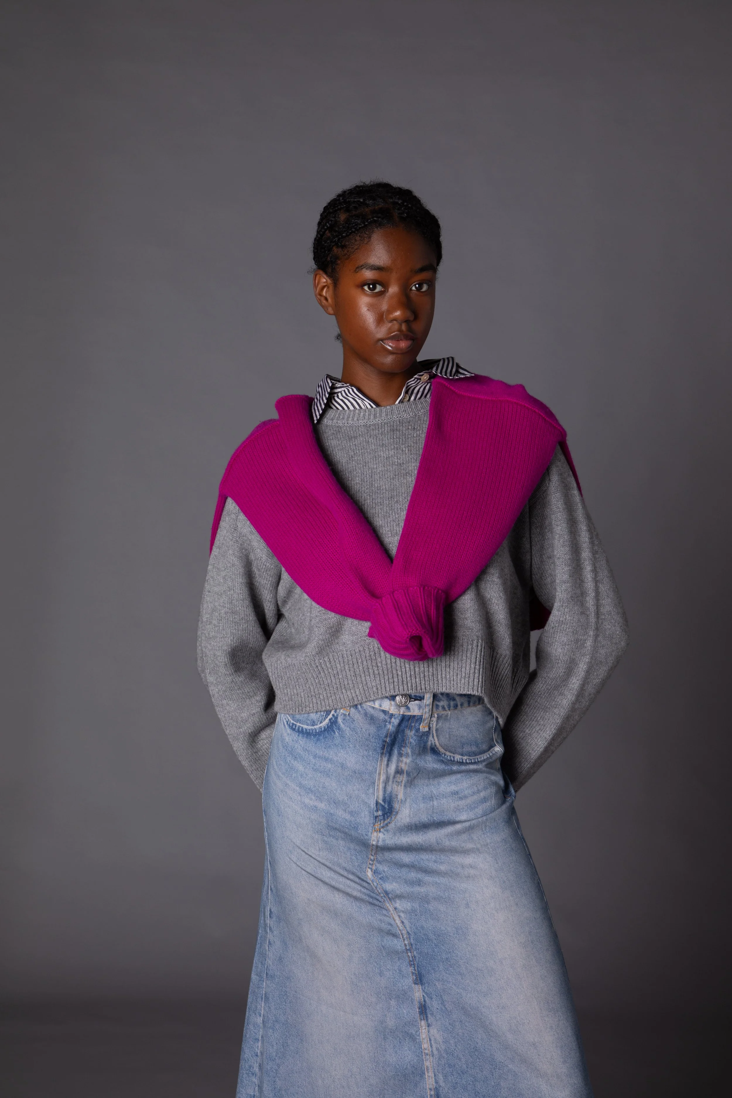 A young woman with dark skin and braided hair stands against a gray background, wearing a gray sweater, a striped shirt underneath, a bright pink sweater draped over her shoulders, and a long denim skirt.